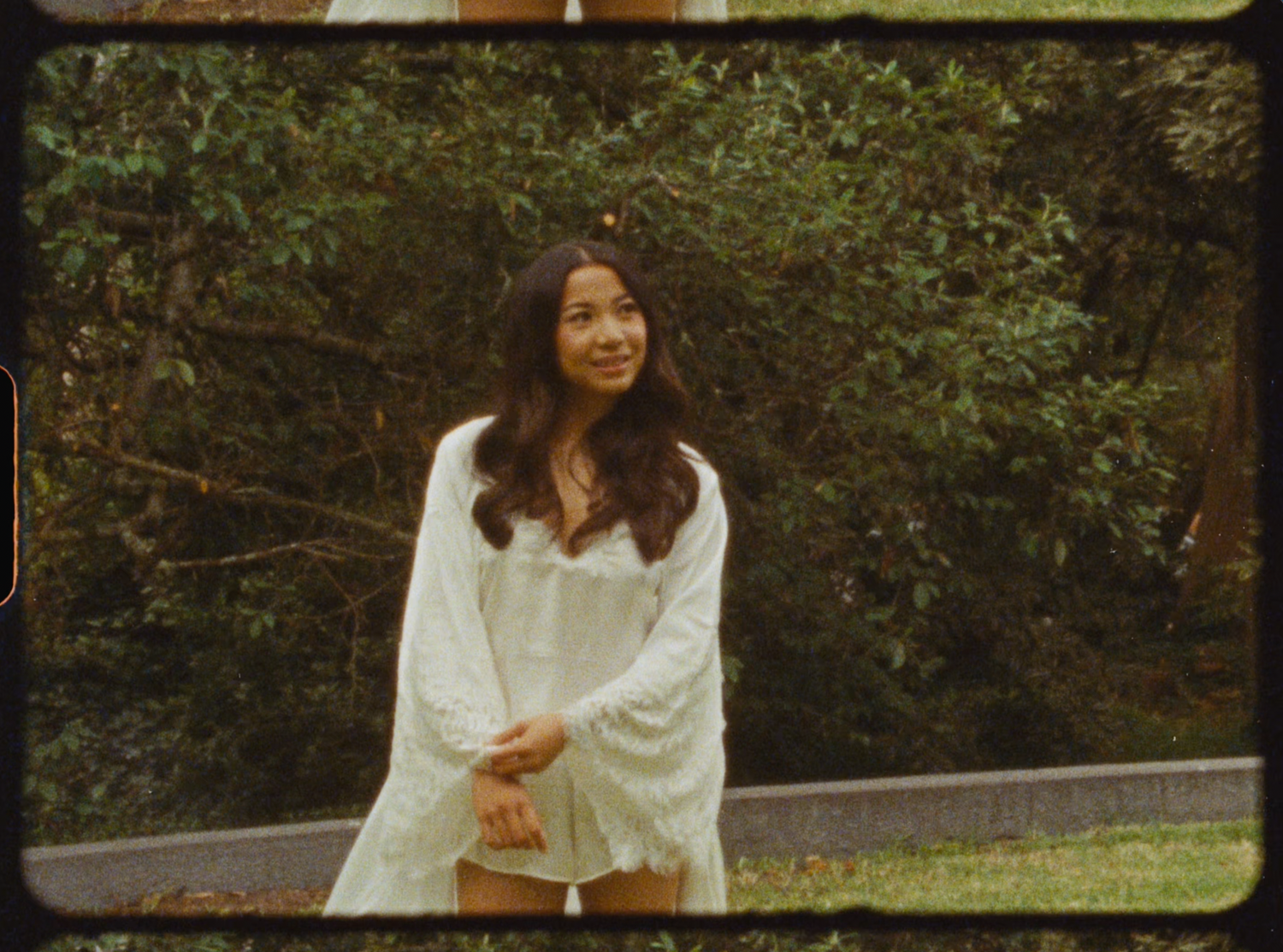 Smiling bride in a white lace robe standing outdoors surrounded by lush green bushes.