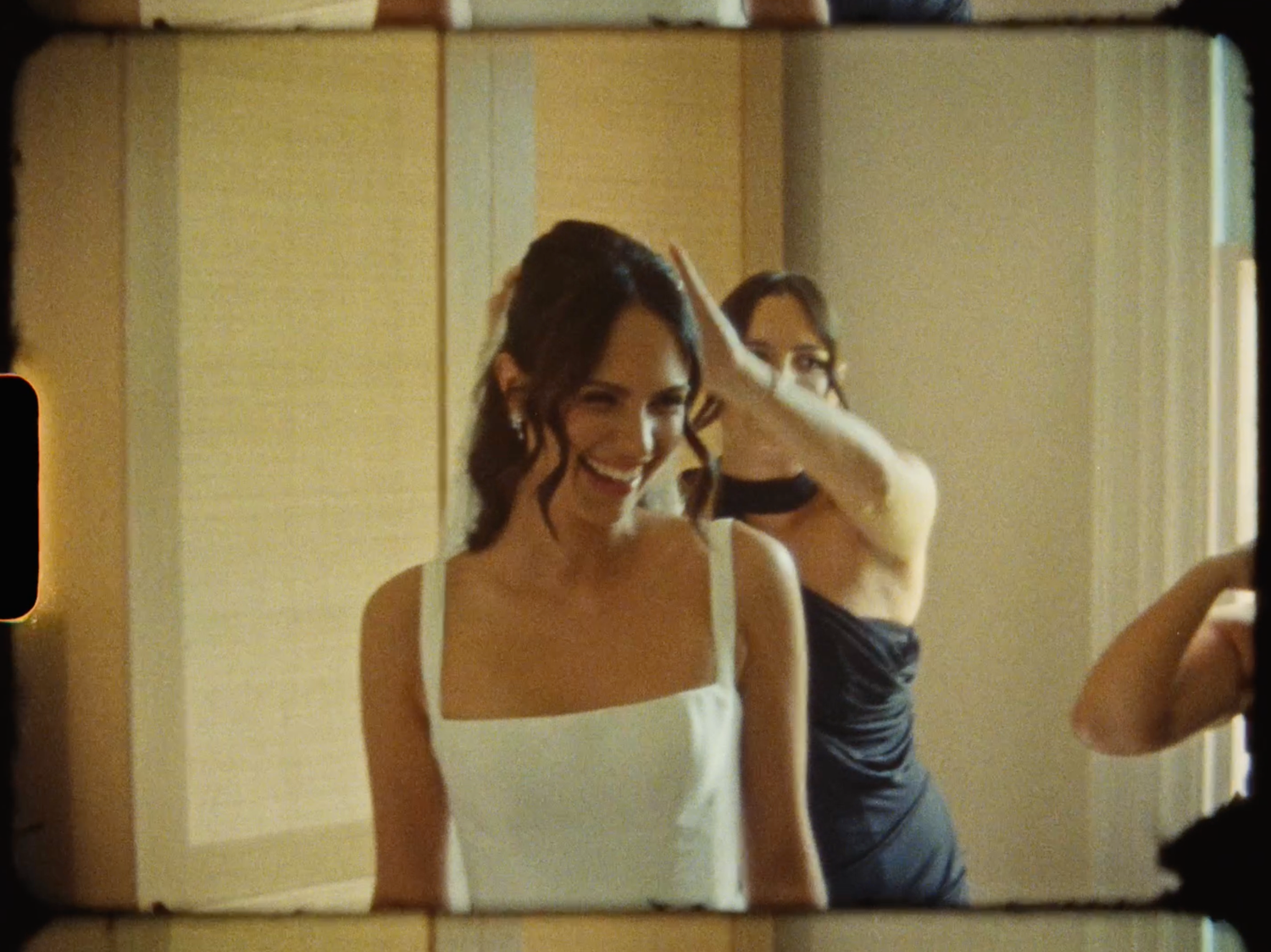 Bride in white gown smiles softly as a bridesmaid adjusts her hair in a sunlit room.