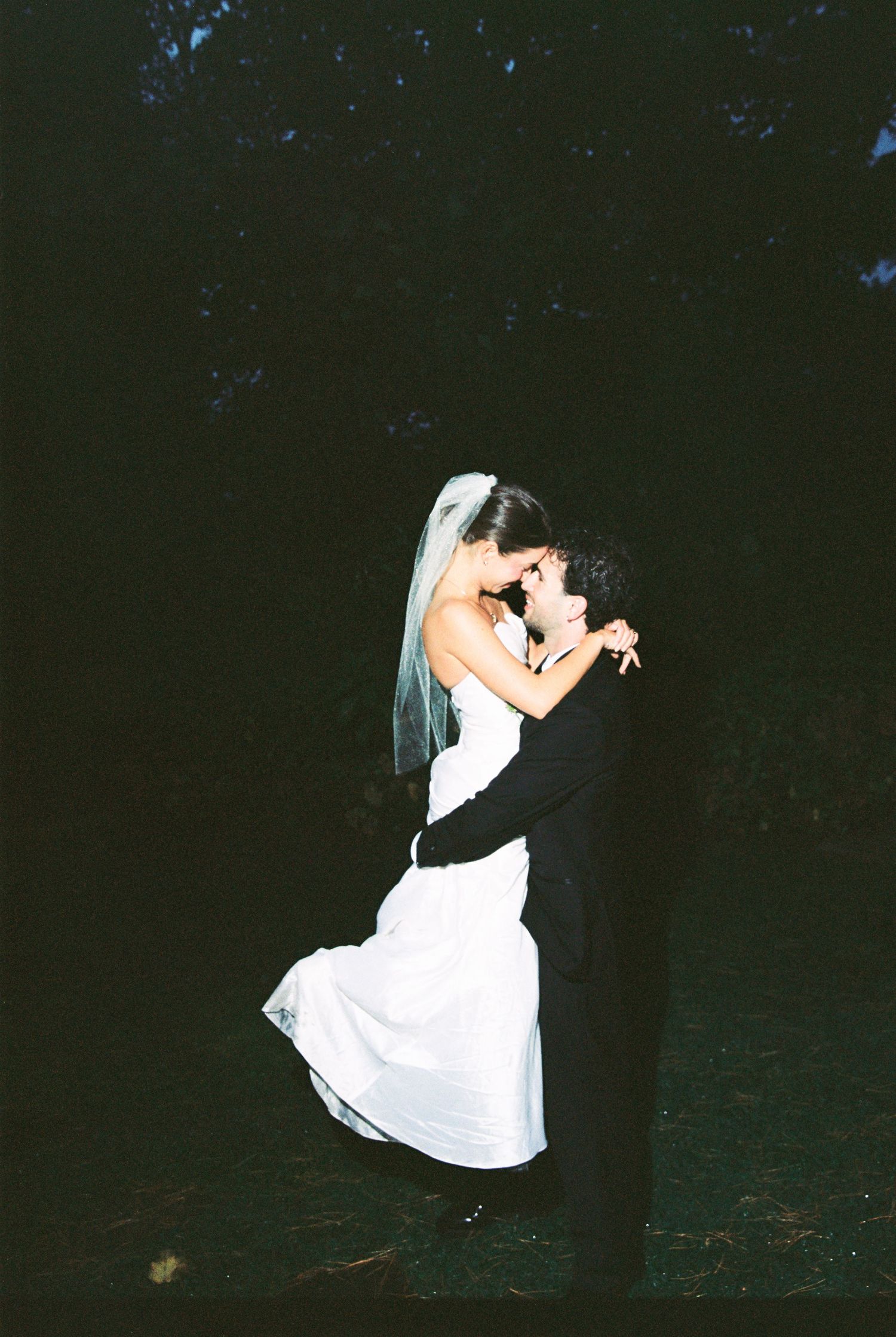 Groom lifting bride in white gown and veil for romantic dance in dark outdoor wedding setting.