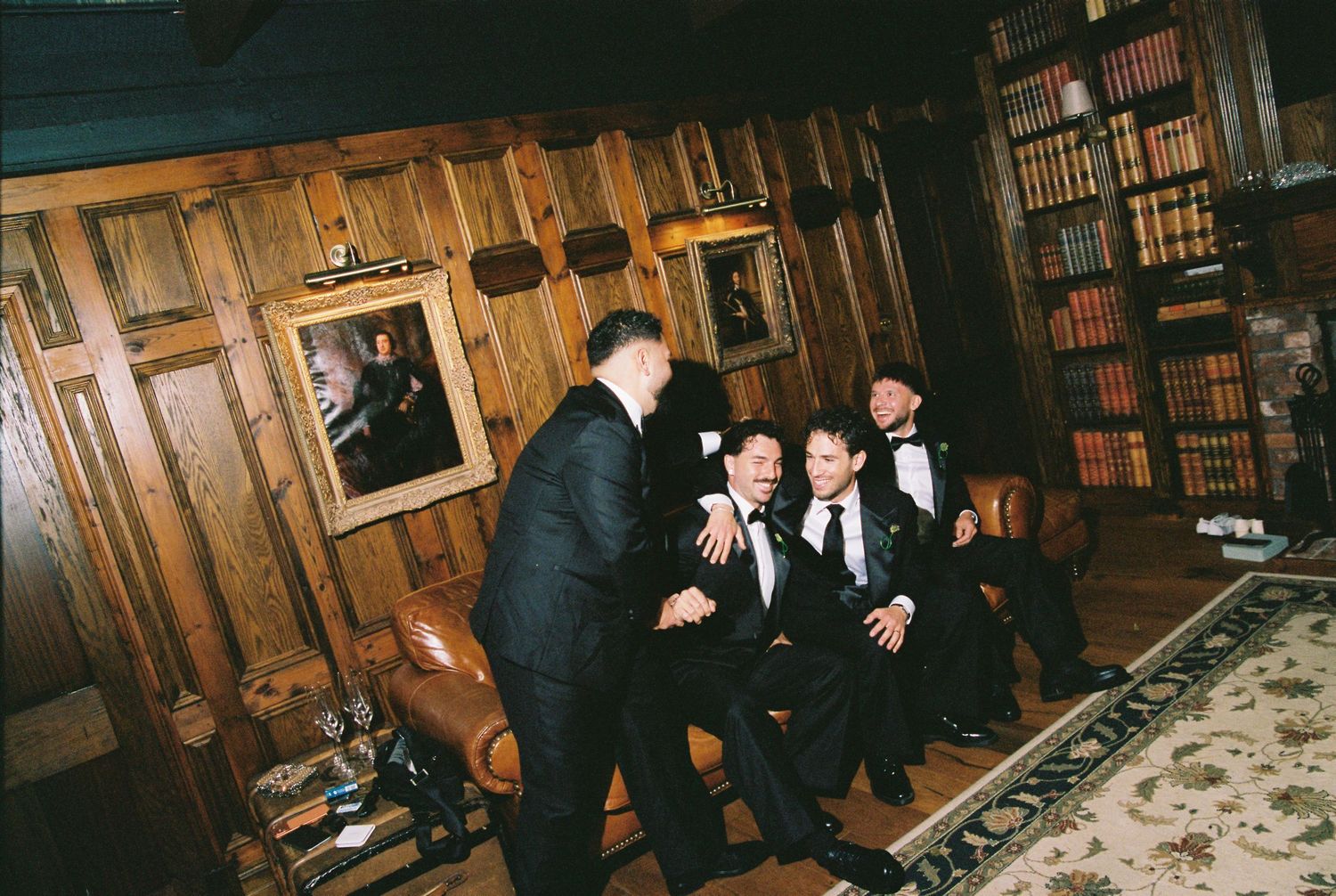Groomsmen in tuxedos laughing together in a wood-paneled library room.