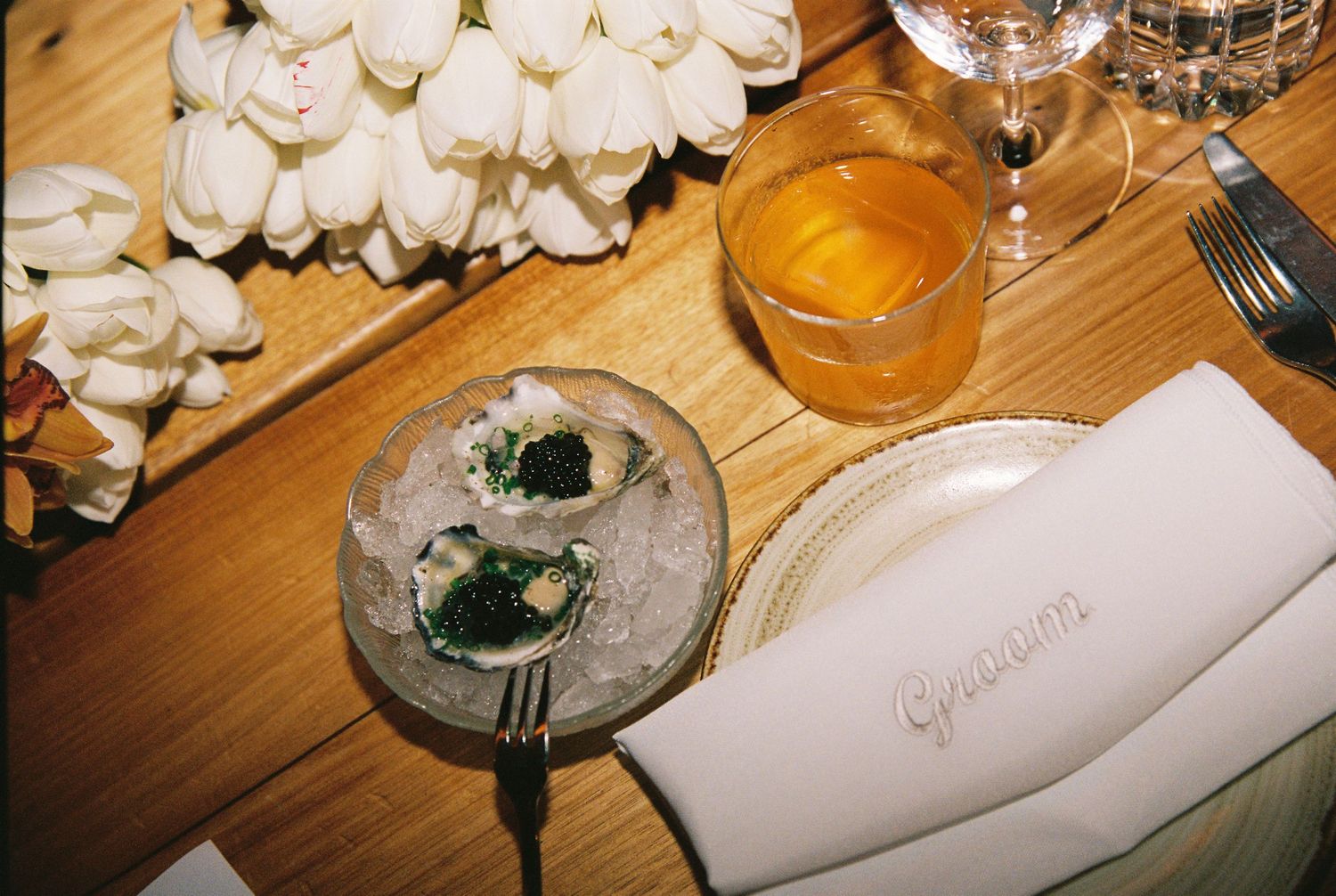 Elegant wedding table setting with caviar appetizers on ice, cocktail drink, and embroidered napkin on wooden table.
