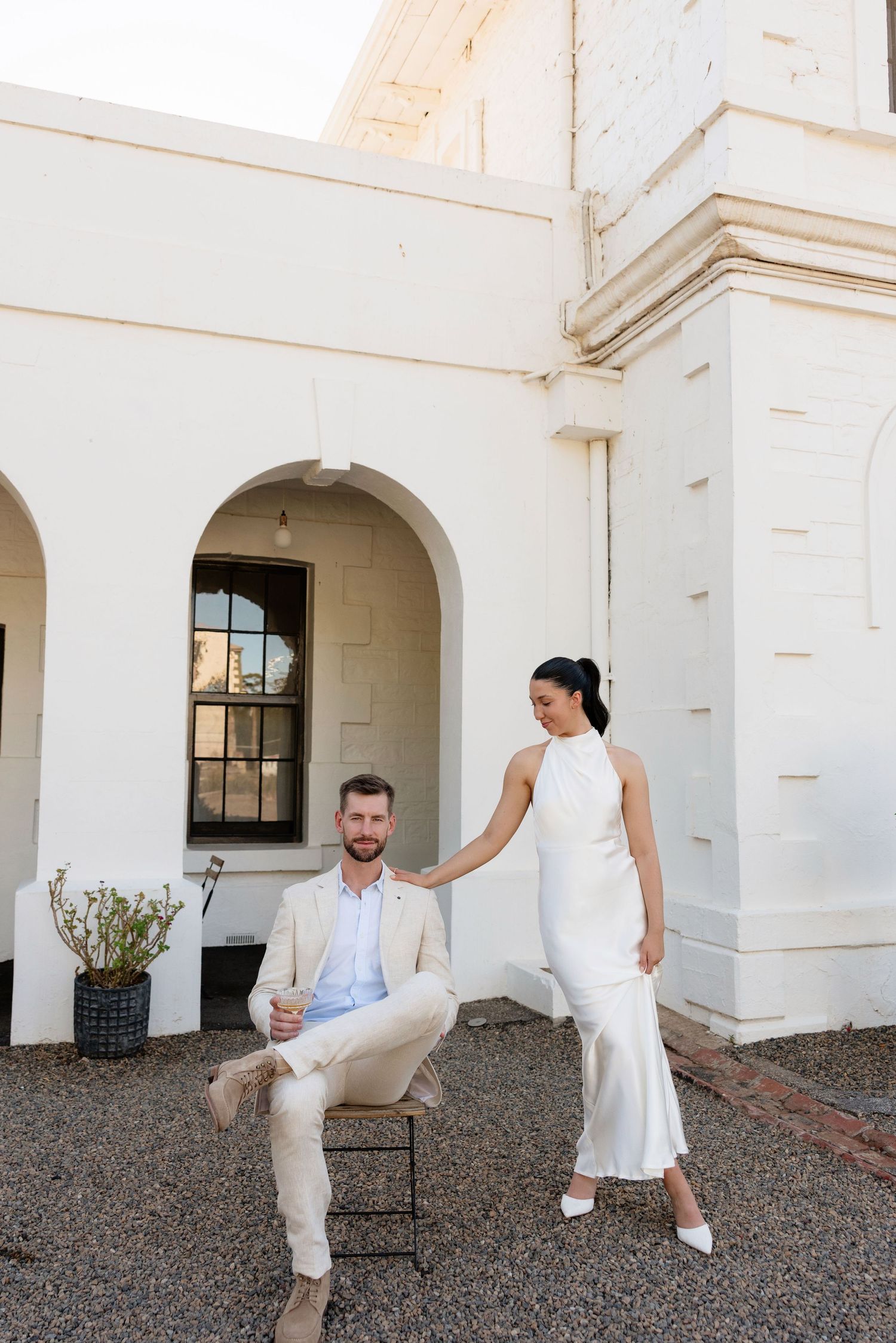 Bride in white halter gown holds grooms hand as he sits outside historic white building.