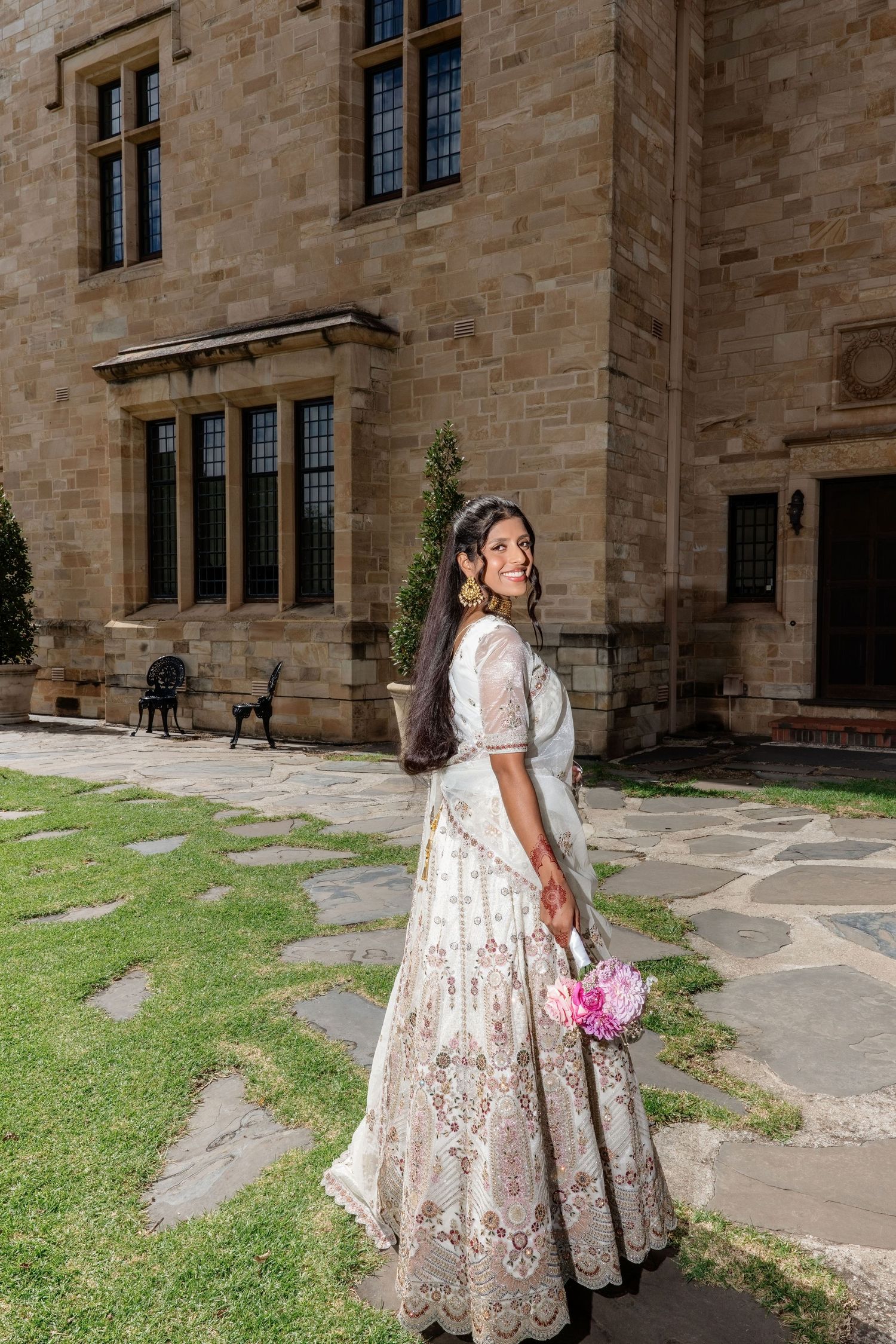 Indian bride in embroidered white lehenga holding pink bouquet, smiling outside historic stone building.