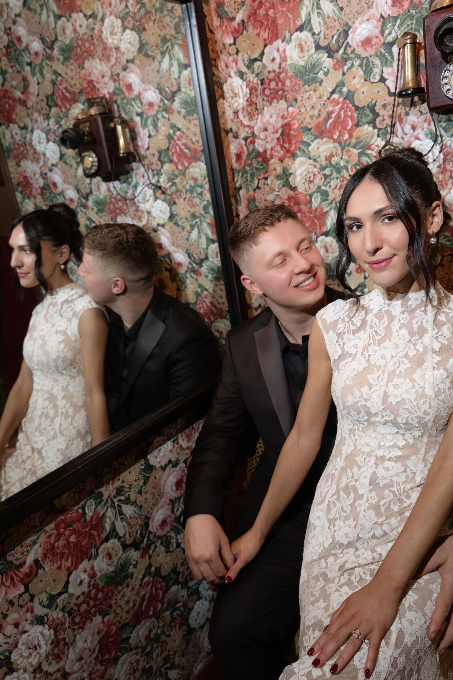 Couple in wedding attire posing in front of floral wallpaper, their reflection visible in a nearby mirror.