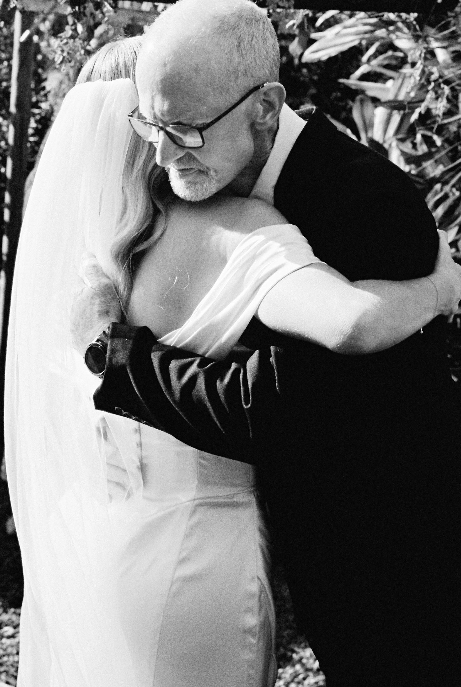 Bride and groom share an emotional embrace, with bride in white gown and veil, groom in black suit.