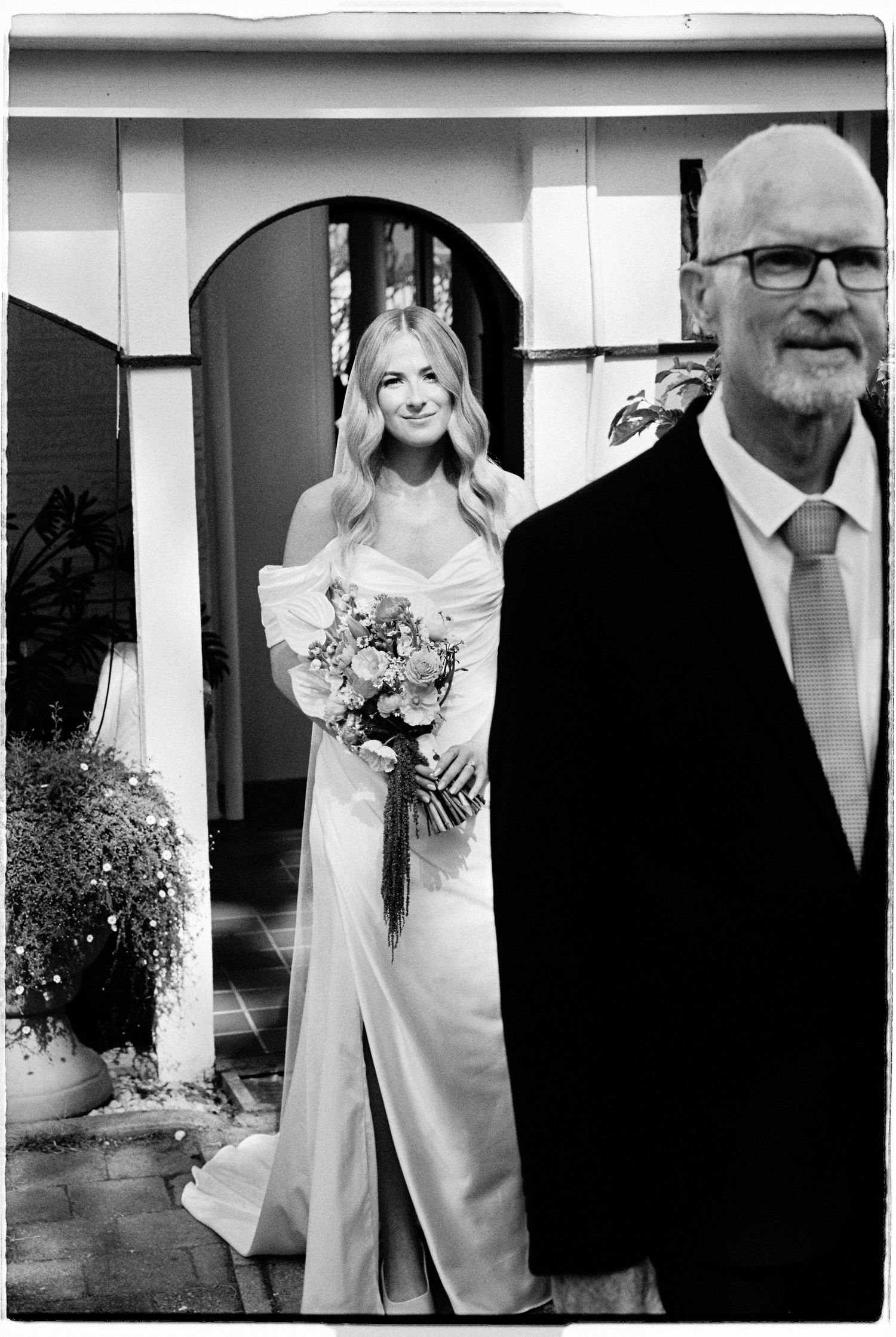 Smiling bride in off-shoulder gown holding bouquet stands with father before wedding ceremony, black and white.