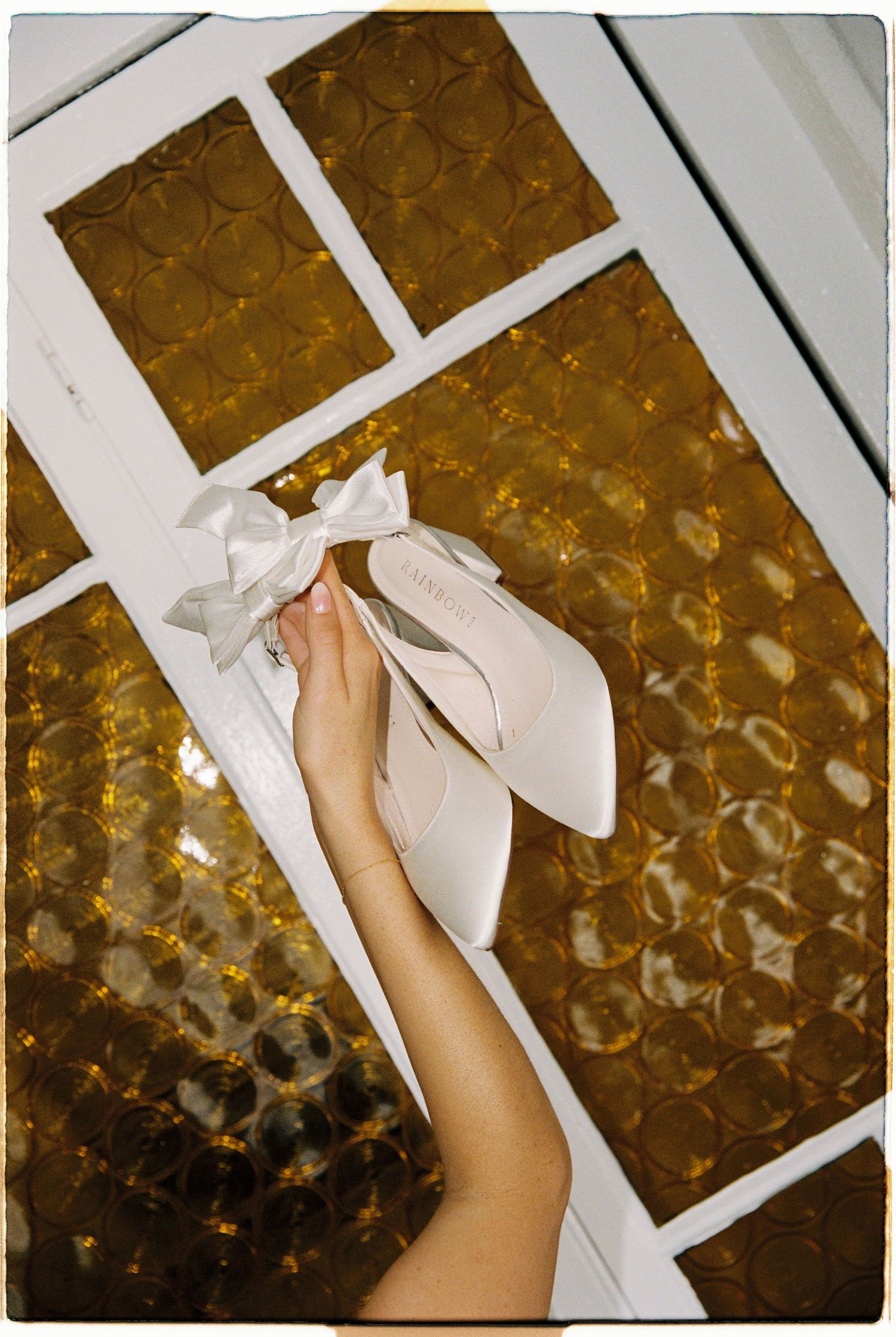 A hand holds a white satin pointed-toe heel with an elegant bow against a golden glass window backdrop.
