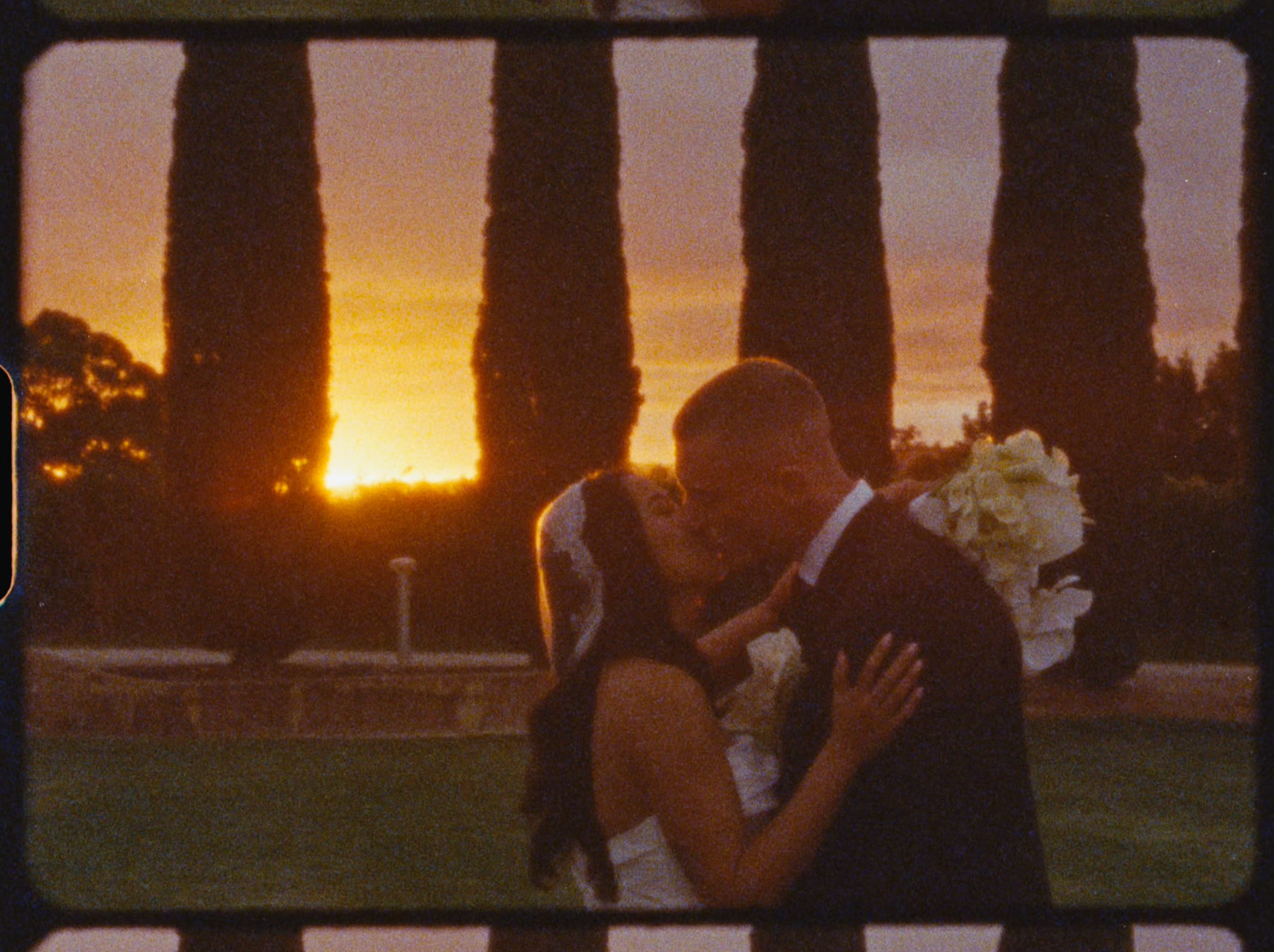 A couple shares a romantic kiss at sunset with tall cypress trees and golden sky backdrop at an outdoor wedding.