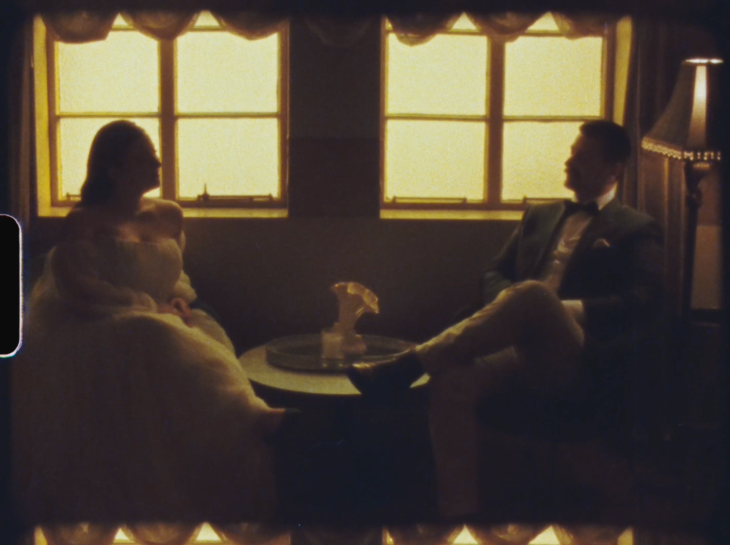Elegant couple in formal attire seated at a small table in a warmly lit vintage setting.