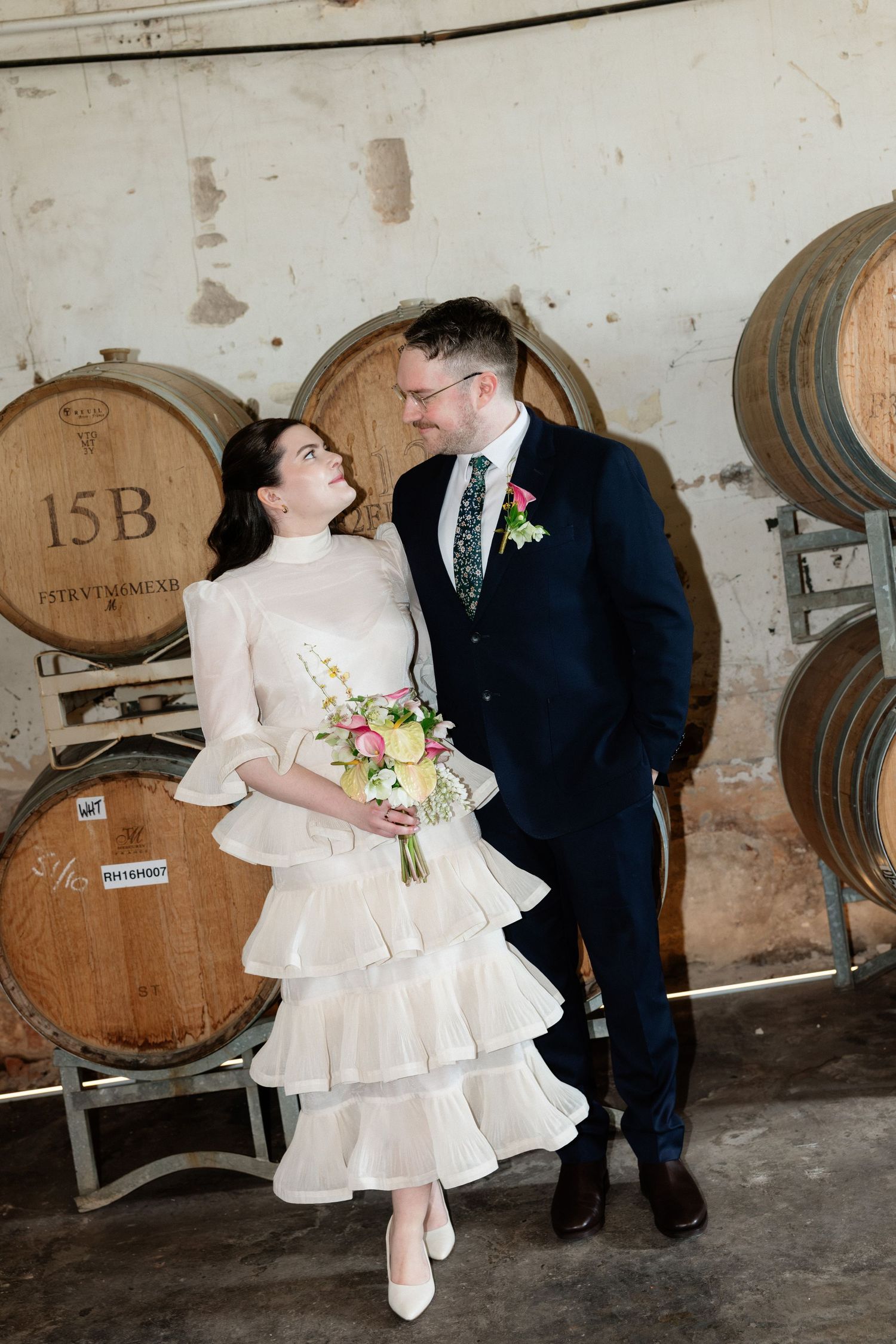 Couple shares romantic glance surrounded by oak barrels in winery wedding setting.