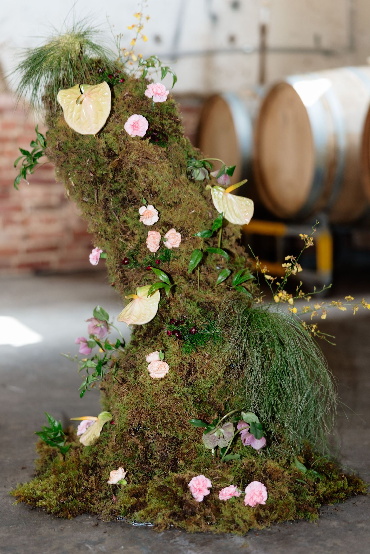 Moss-covered sculptural installation adorned with pink flowers and anthurium leaves displayed in a rustic winery setting.