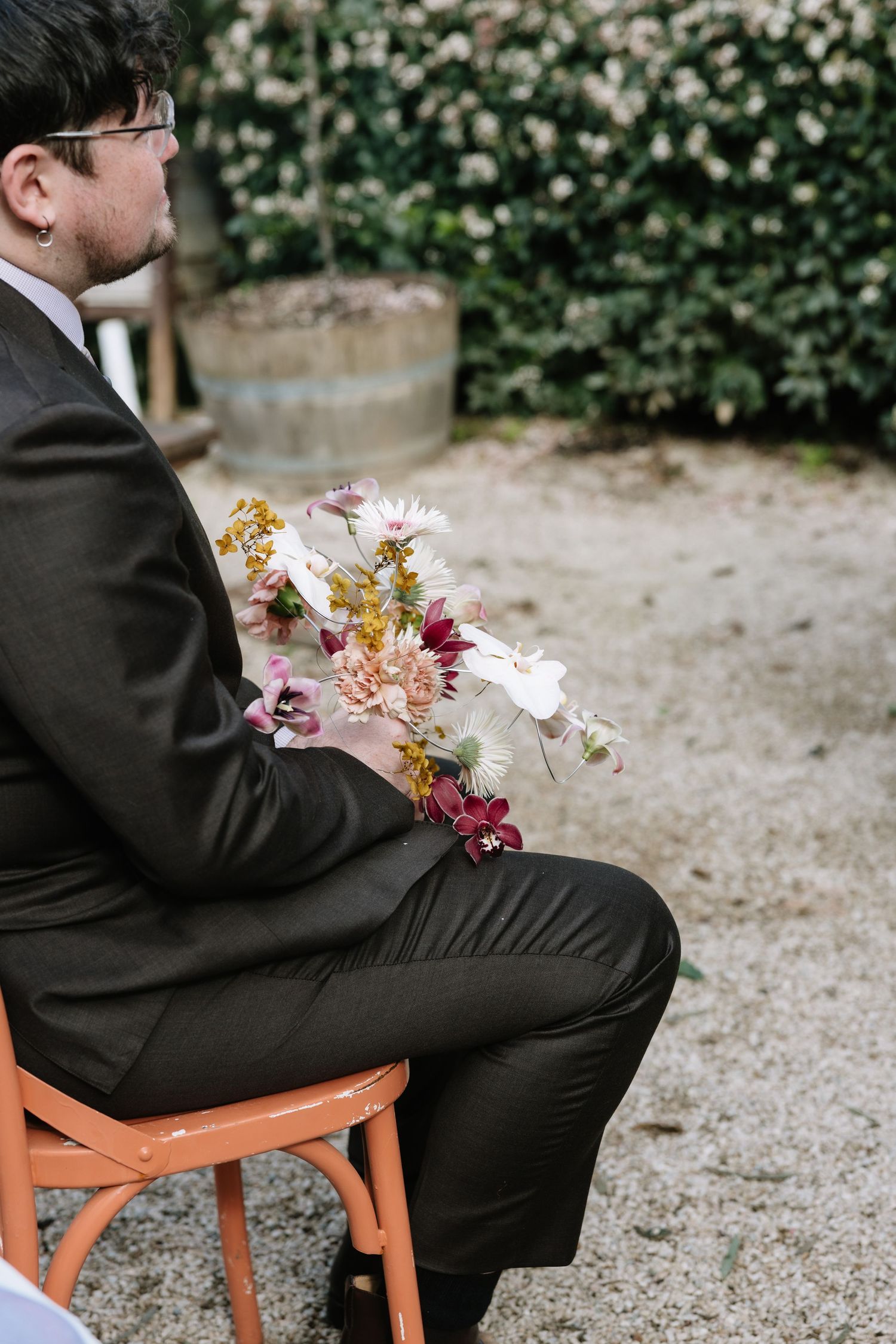 Man in dark suit sitting on orange chair holding a colorful bridal bouquet with pink and burgundy flowers outdoors.