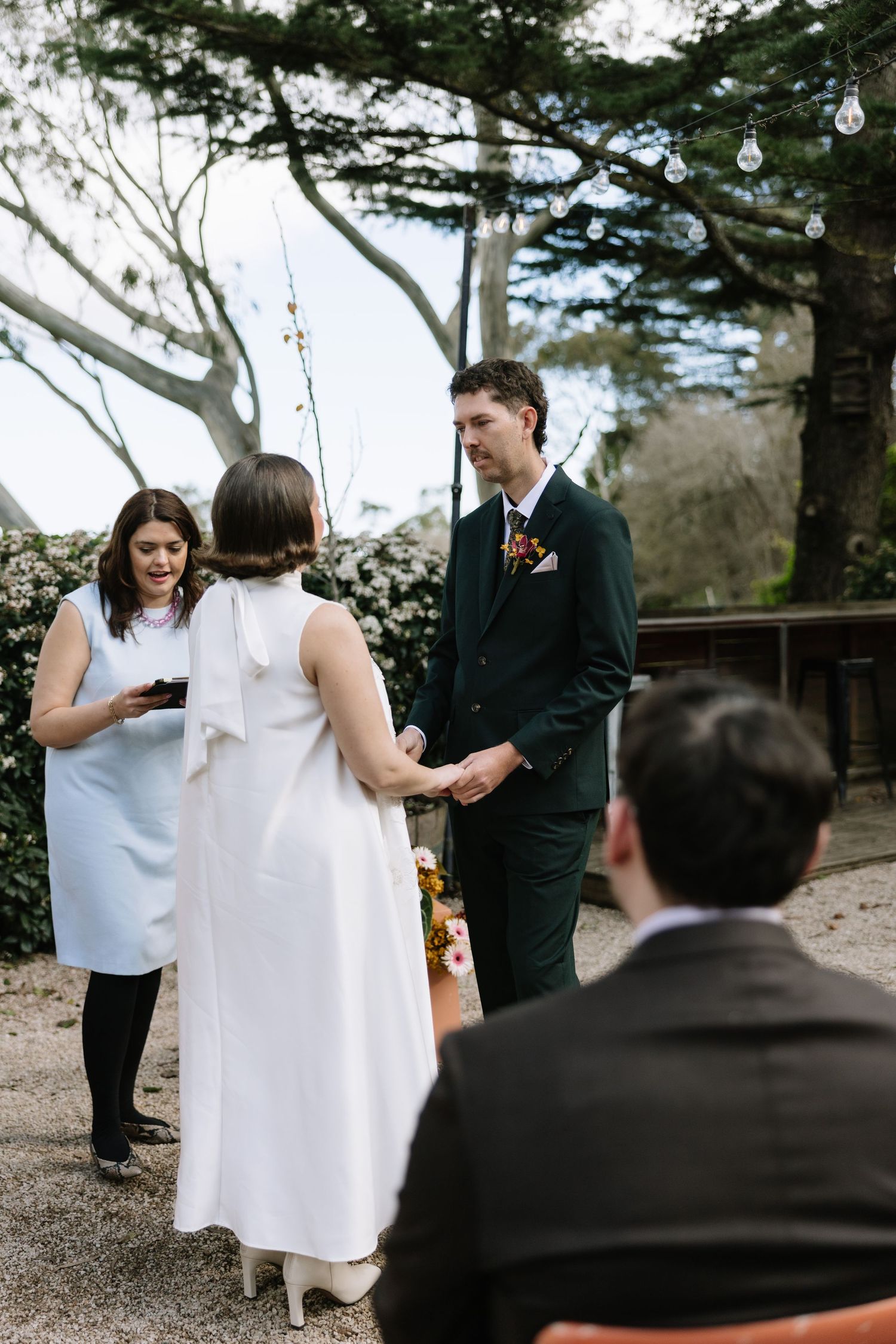 Outdoor wedding ceremony with bride in white dress, groom in dark green suit holding hands, officiant reading vows.