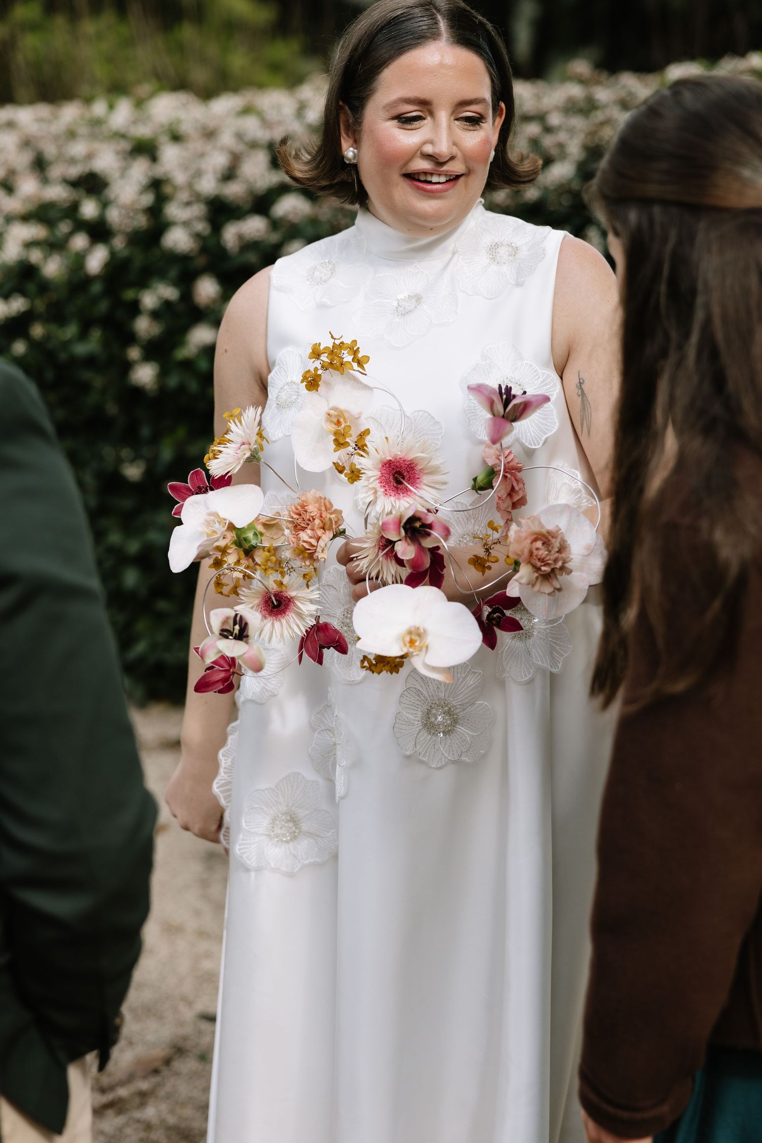 Bride in white floral gown holding colorful orchid bouquet, smiling at outdoor wedding ceremony.