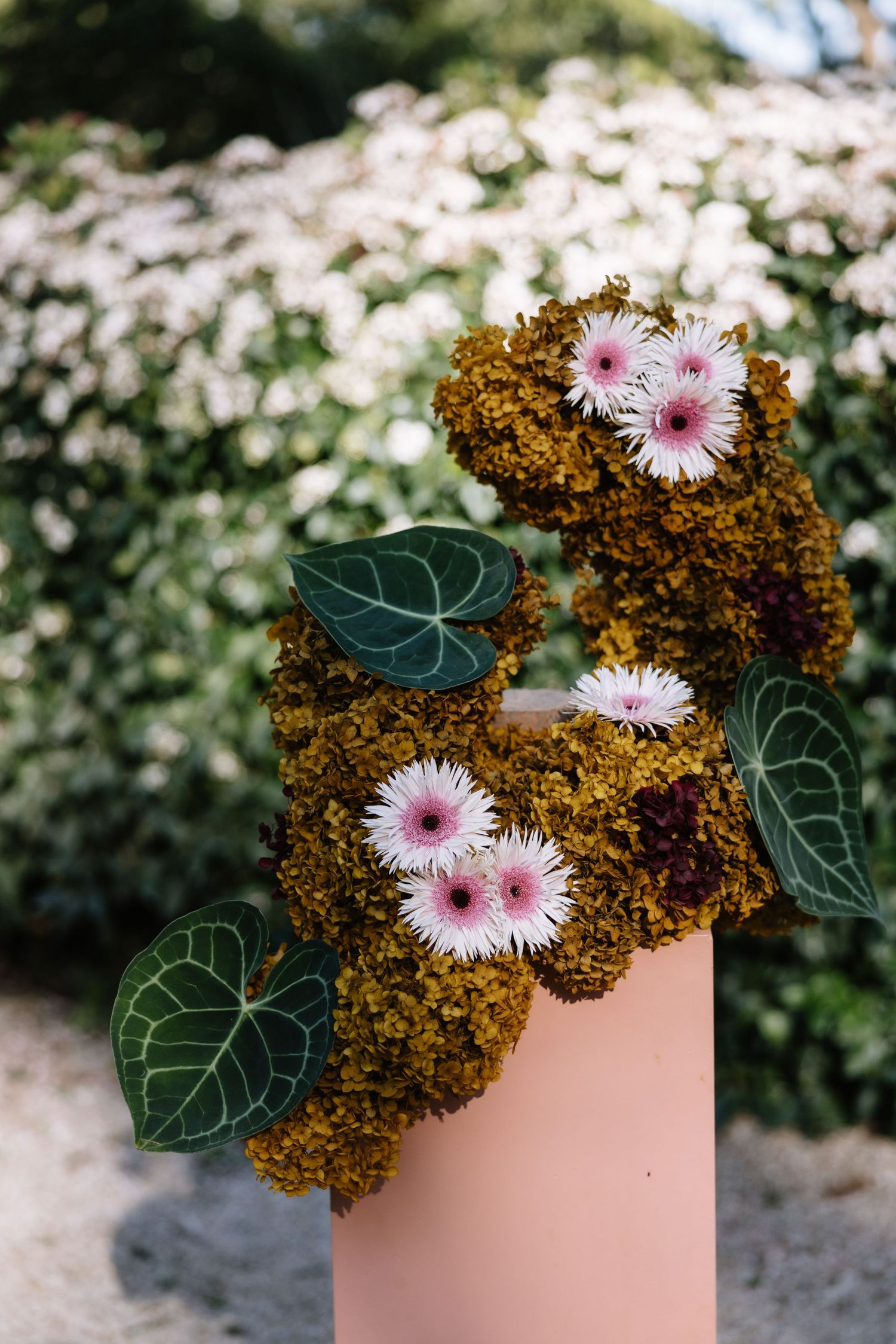 Floral arrangement with pink gerbera daisies and green leaves on a pink pedestal, set against a lush garden backdrop.