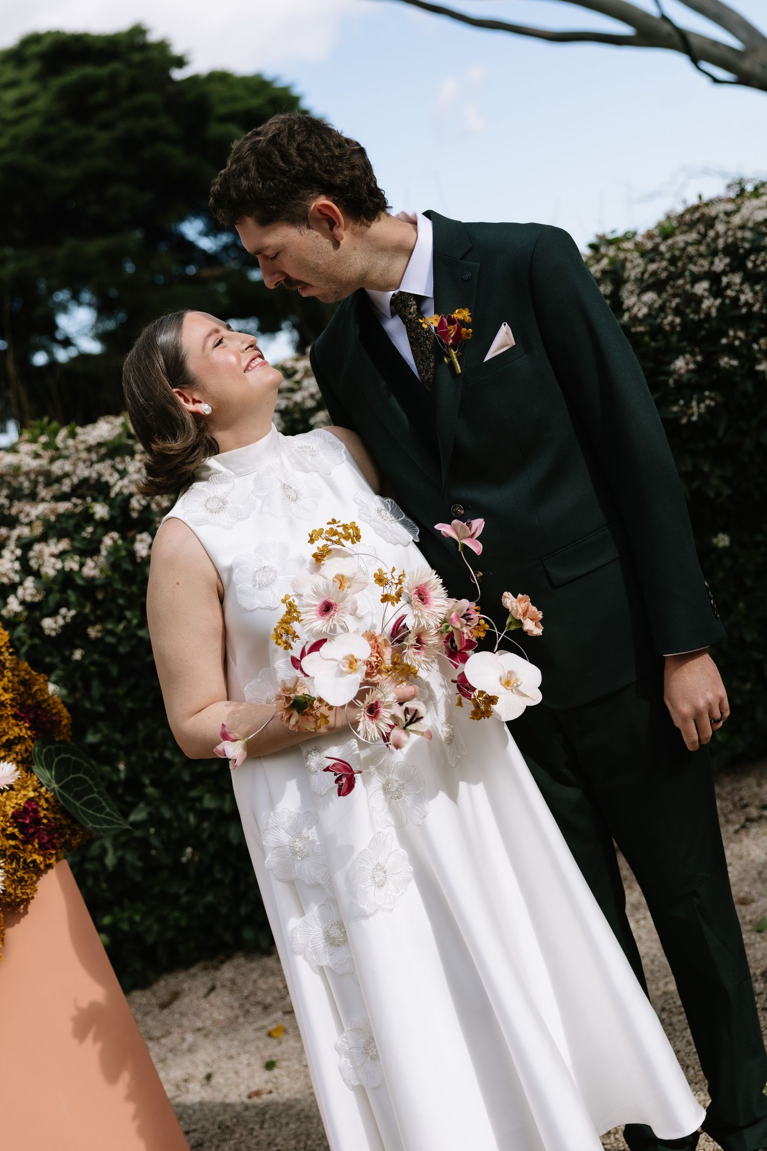 Bride in white floral gown holds orchid bouquet, smiling at groom in dark suit with boutonniere outdoors.