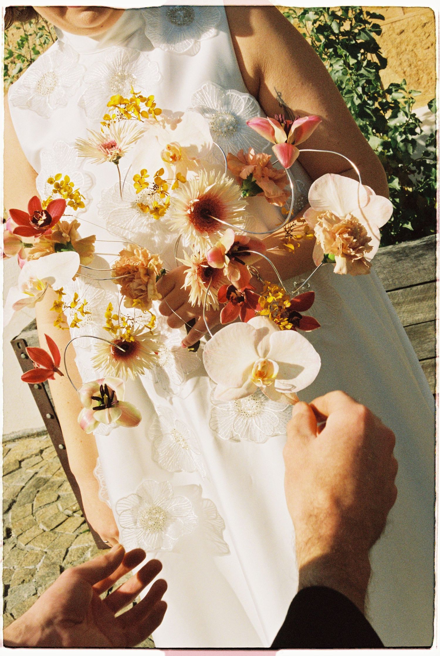 Bride holding a colorful wedding bouquet with white orchids, red and yellow blooms, while a hand adjusts the arrangement.