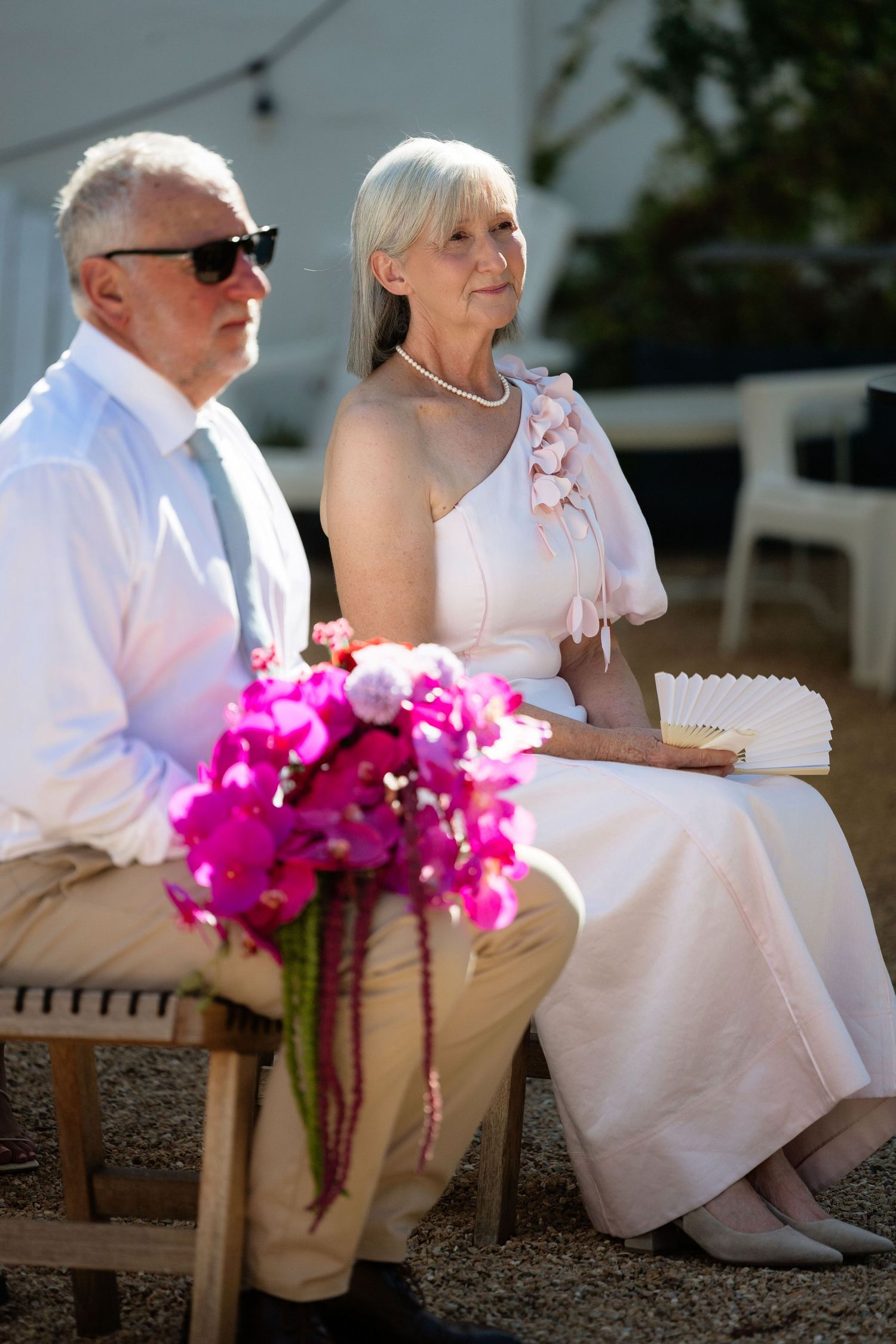 Elegant senior couple seated outdoors at a wedding ceremony, woman in white dress holding pink flowers bouquet.