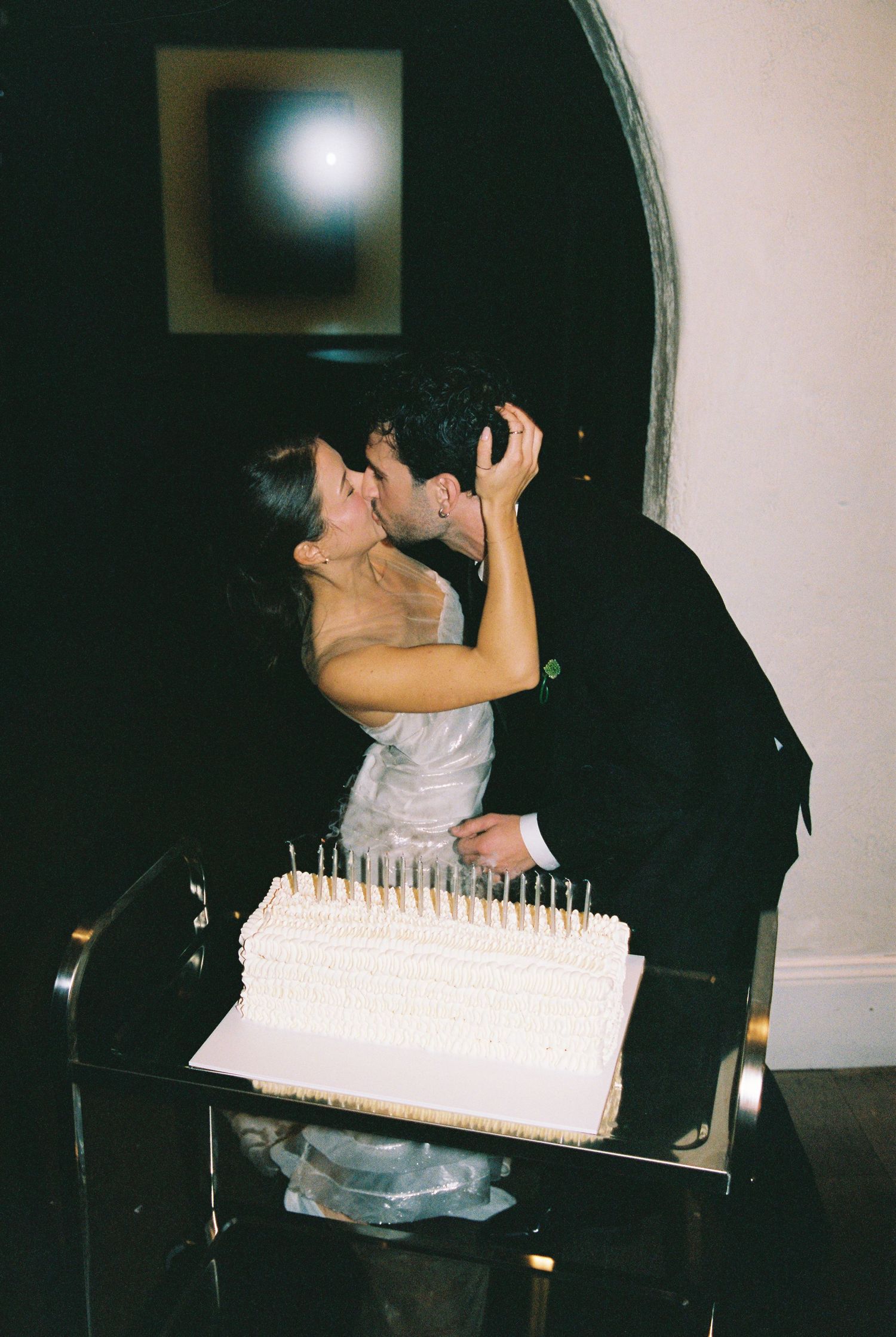 Bride and groom sharing a kiss behind their white wedding cake with candles.