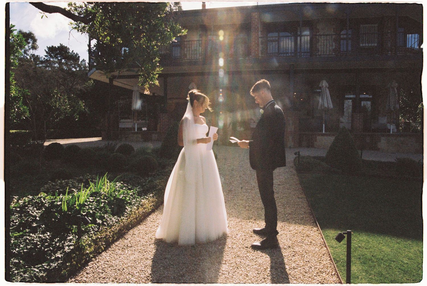 Bride and groom exchange vows in sunlit garden with historic building backdrop.