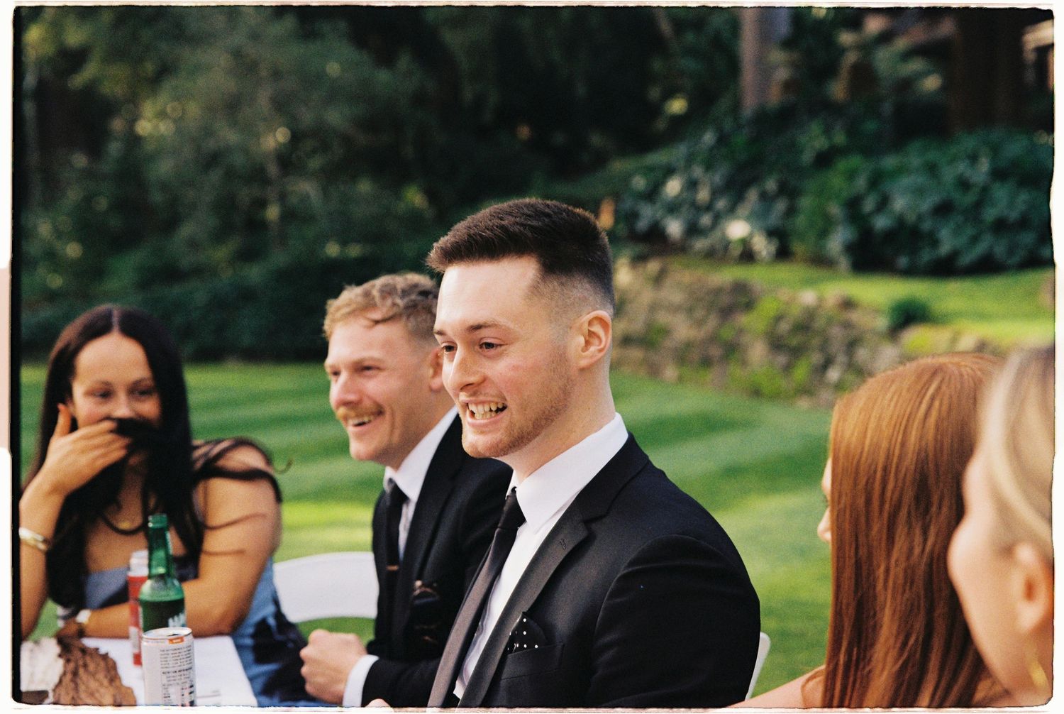 Wedding guests in formal attire smiling and laughing together at an outdoor reception table.