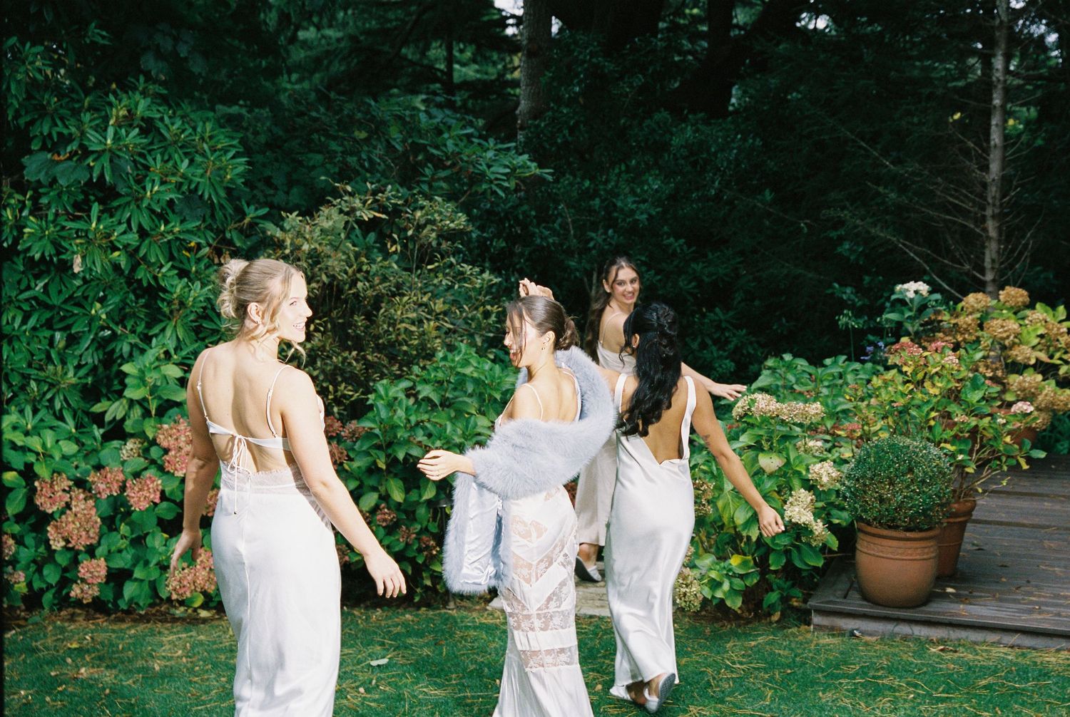 Bridesmaids in white dresses dancing joyfully in a lush garden setting.