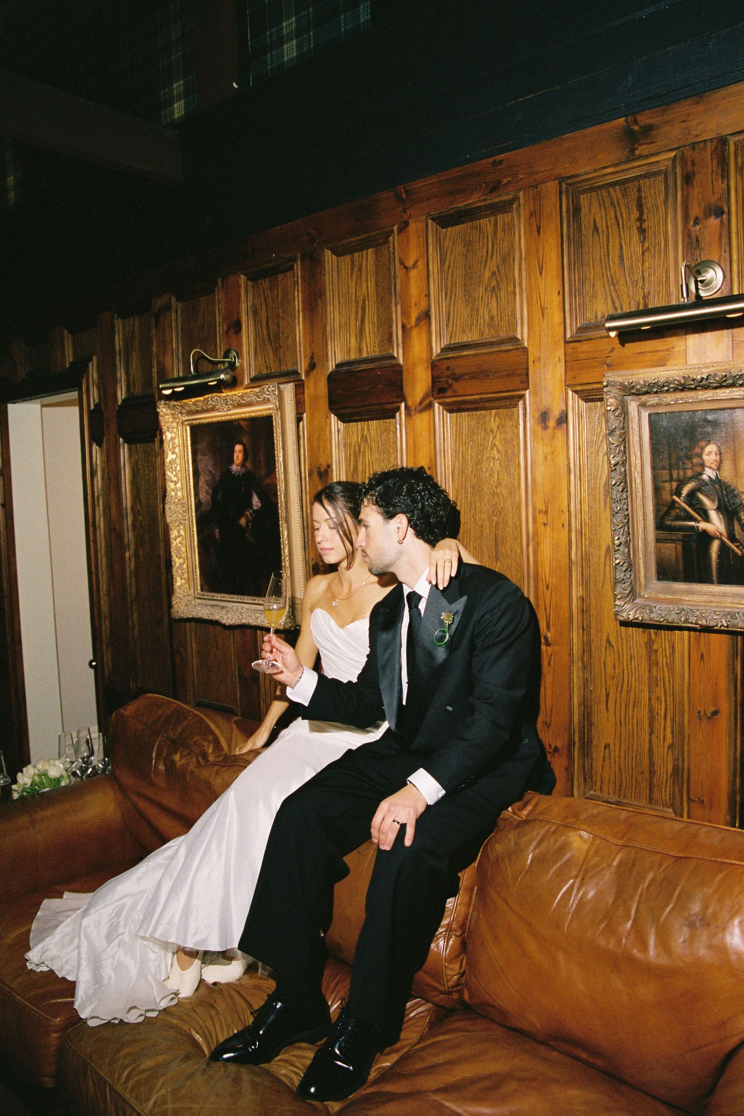Bride and groom sit together on leather sofa surrounded by portrait paintings on wood walls.