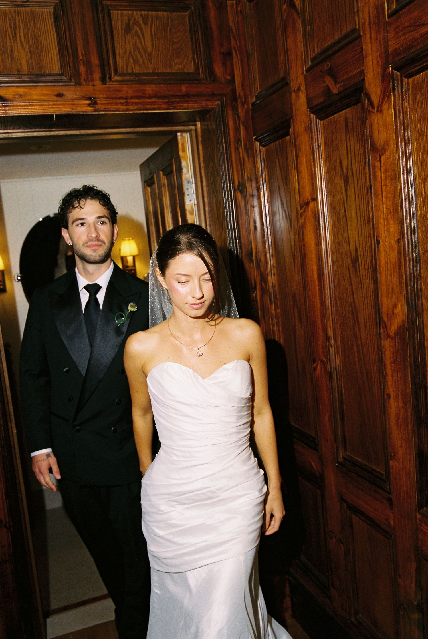 Bride in strapless white wedding gown and groom in black tuxedo walk through grand wooden doors.