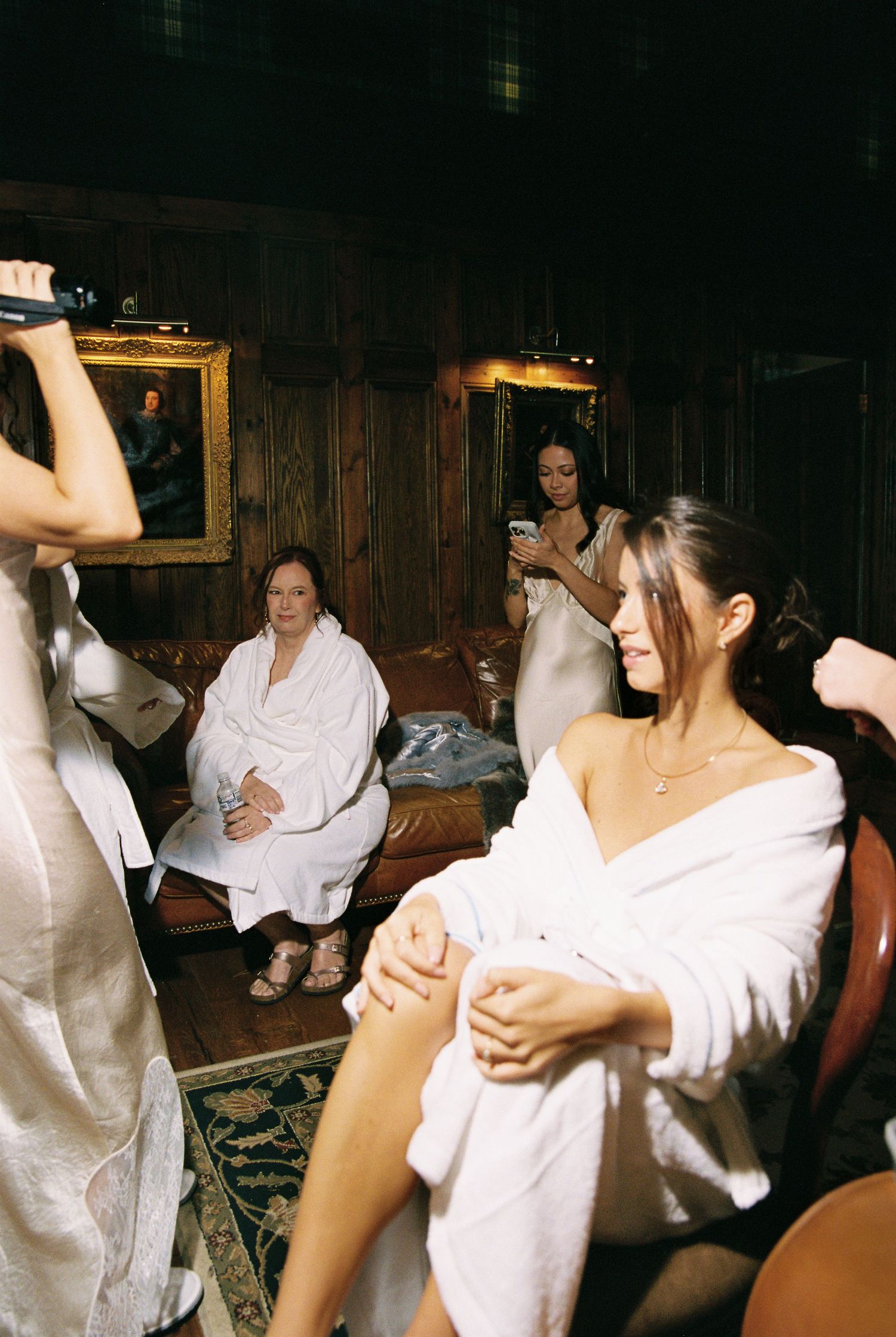 Bride and bridesmaids in white robes preparing for wedding in elegant wood-paneled dressing room.