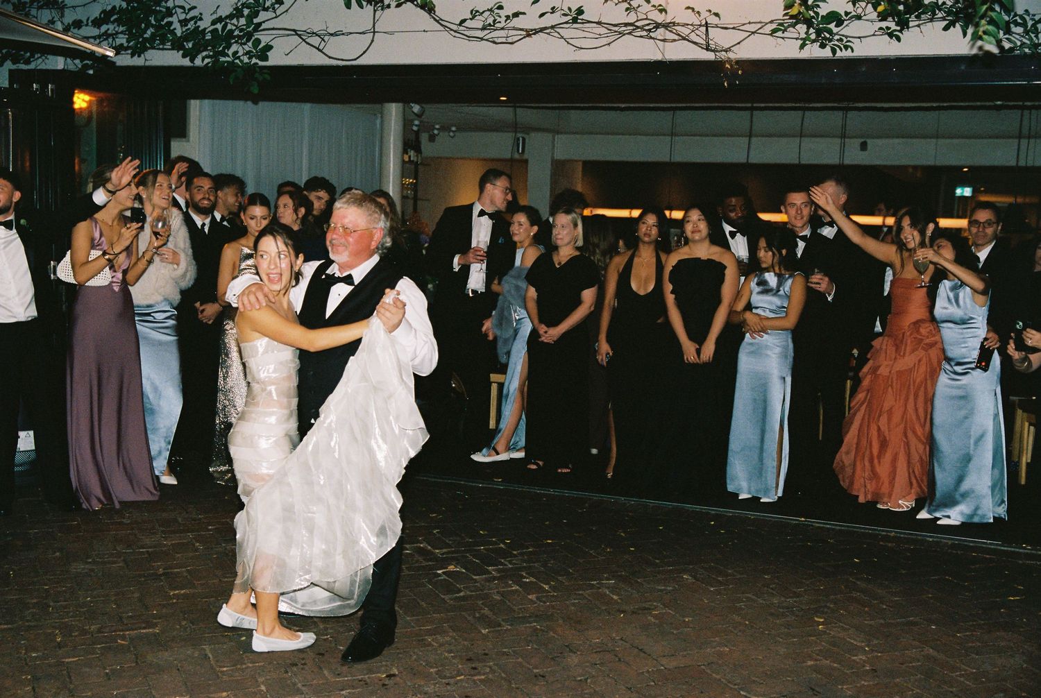 Bride and groom share first dance at wedding reception while guests watch and celebrate outdoors at night.