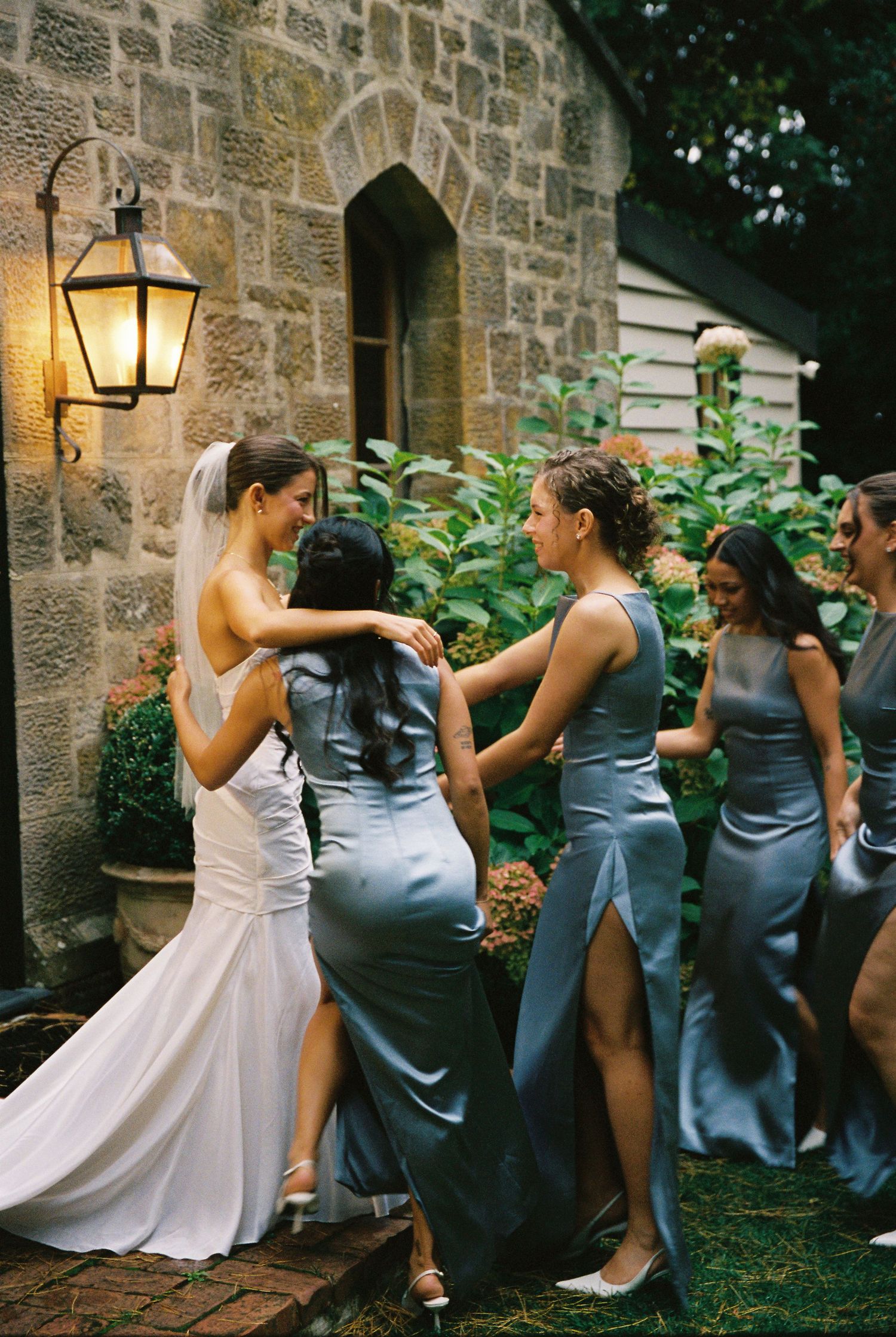 Bride in white gown embracing bridesmaids in blue satin dresses outside stone building at dusk.