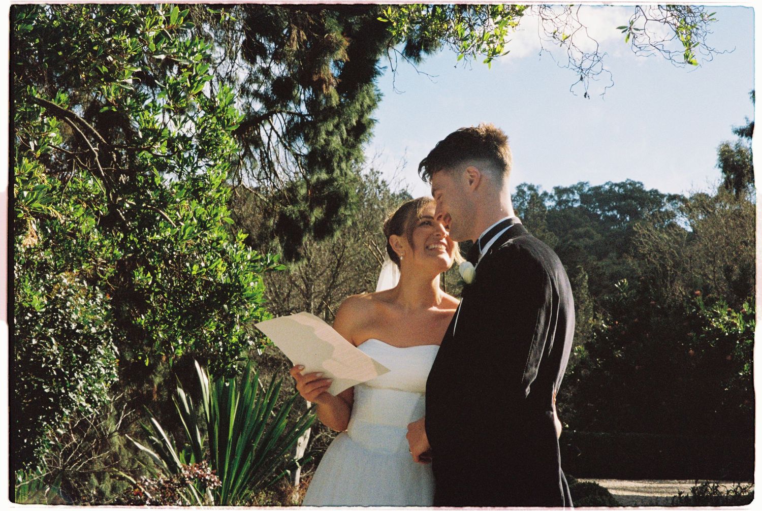 Bride and groom sharing an intimate moment during outdoor wedding ceremony among green trees.