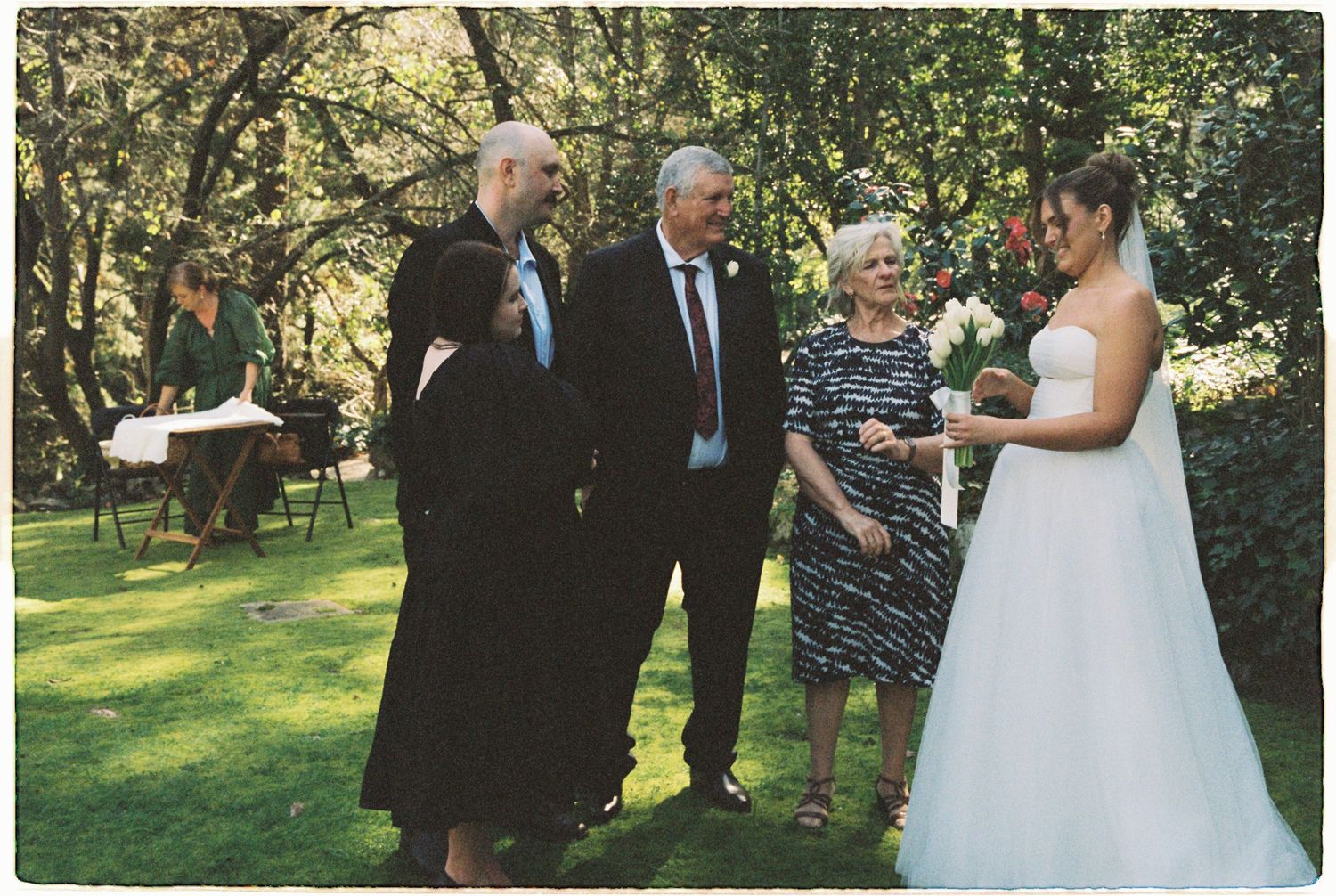 Bride in white gown greets guests including family members during outdoor garden wedding ceremony.