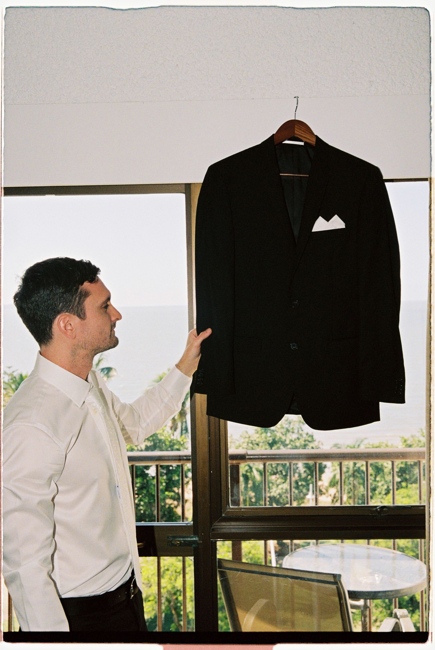 Groom in white shirt pointing at black suit jacket hanging on hanger near balcony window.