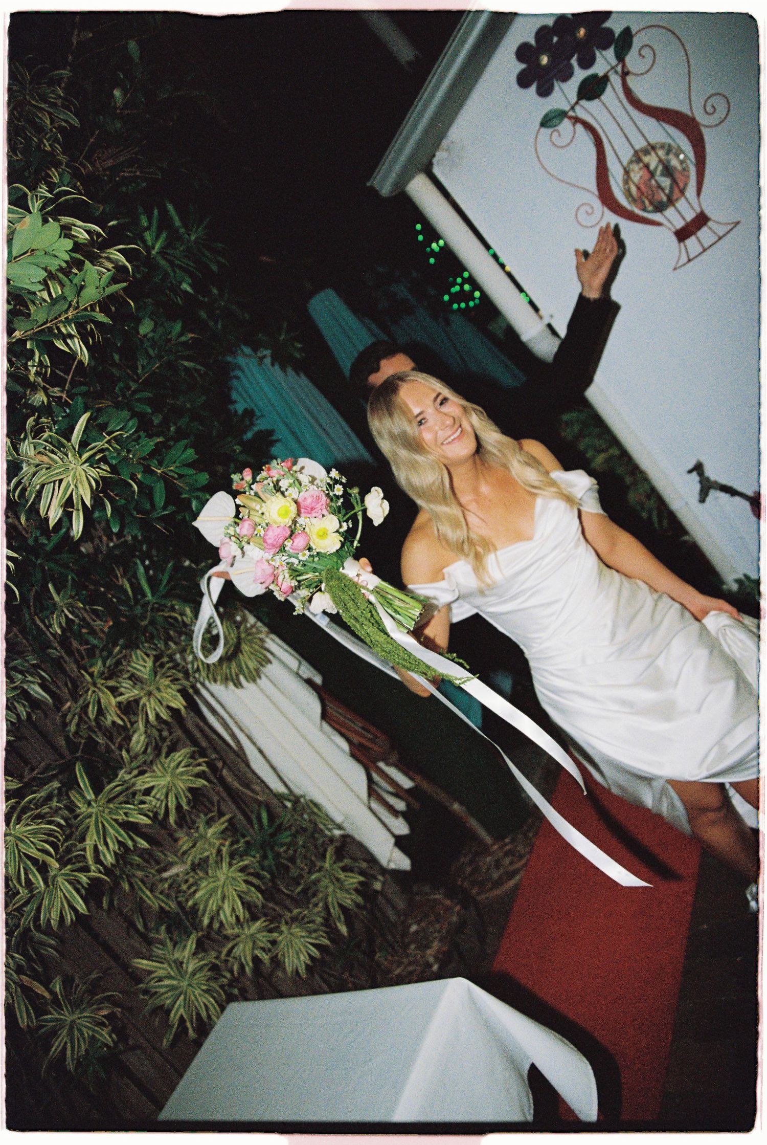 A joyful blonde bride in a white off-shoulder dress holds a ribbon-tied floral bouquet at her wedding venue.