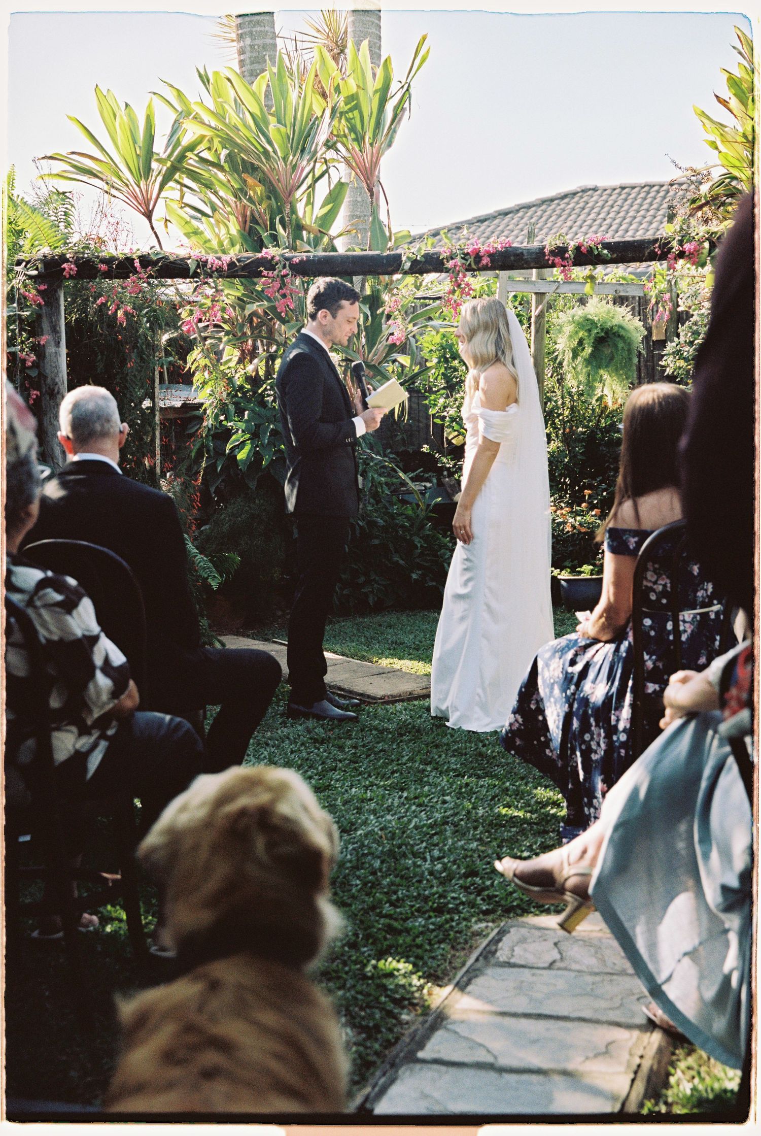Groom reads vows to bride in white dress under floral pergola at intimate garden wedding ceremony.
