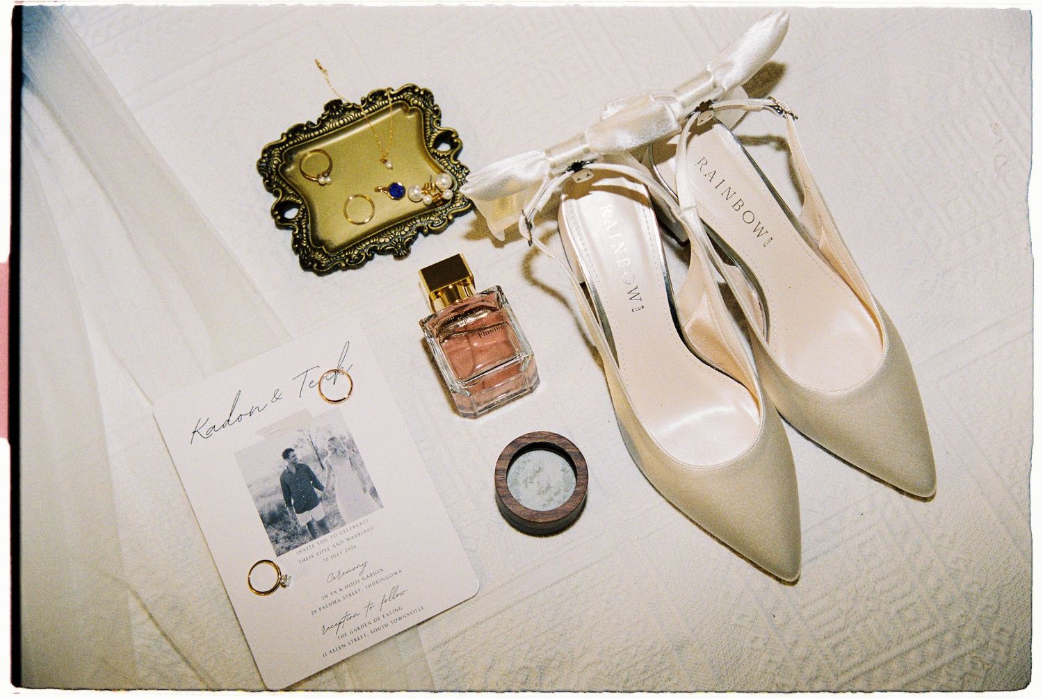 Wedding flat lay featuring nude heels, perfume, gold ring, invitation card, and ribbon accessories on white surface.