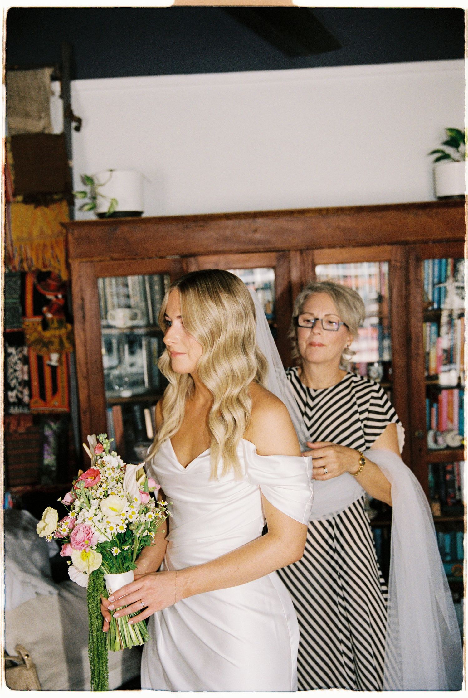 Mother helping bride with veil while she holds floral bouquet near wooden bookcase.