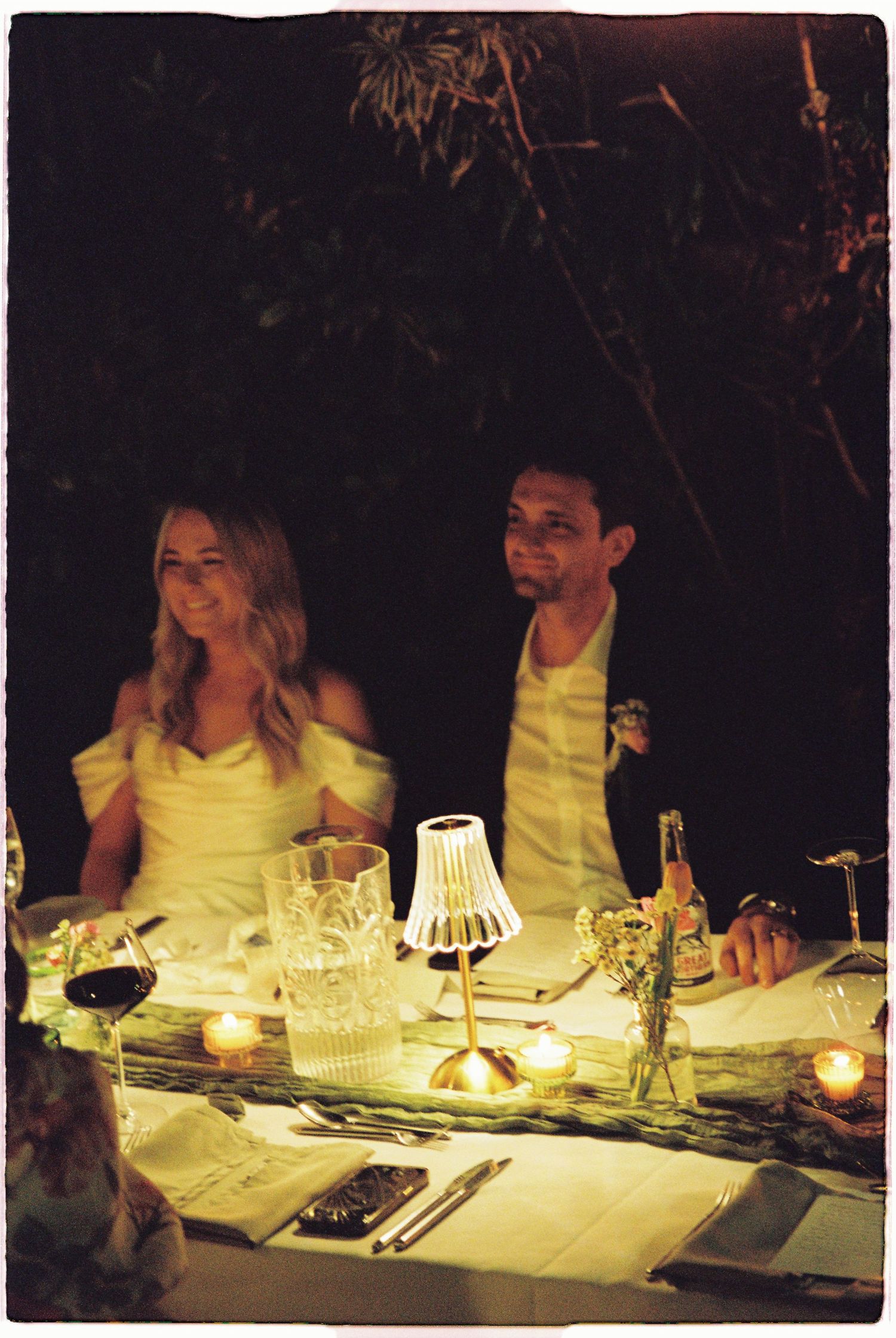 Bride and groom seated at candlelit outdoor wedding reception dinner table at night.