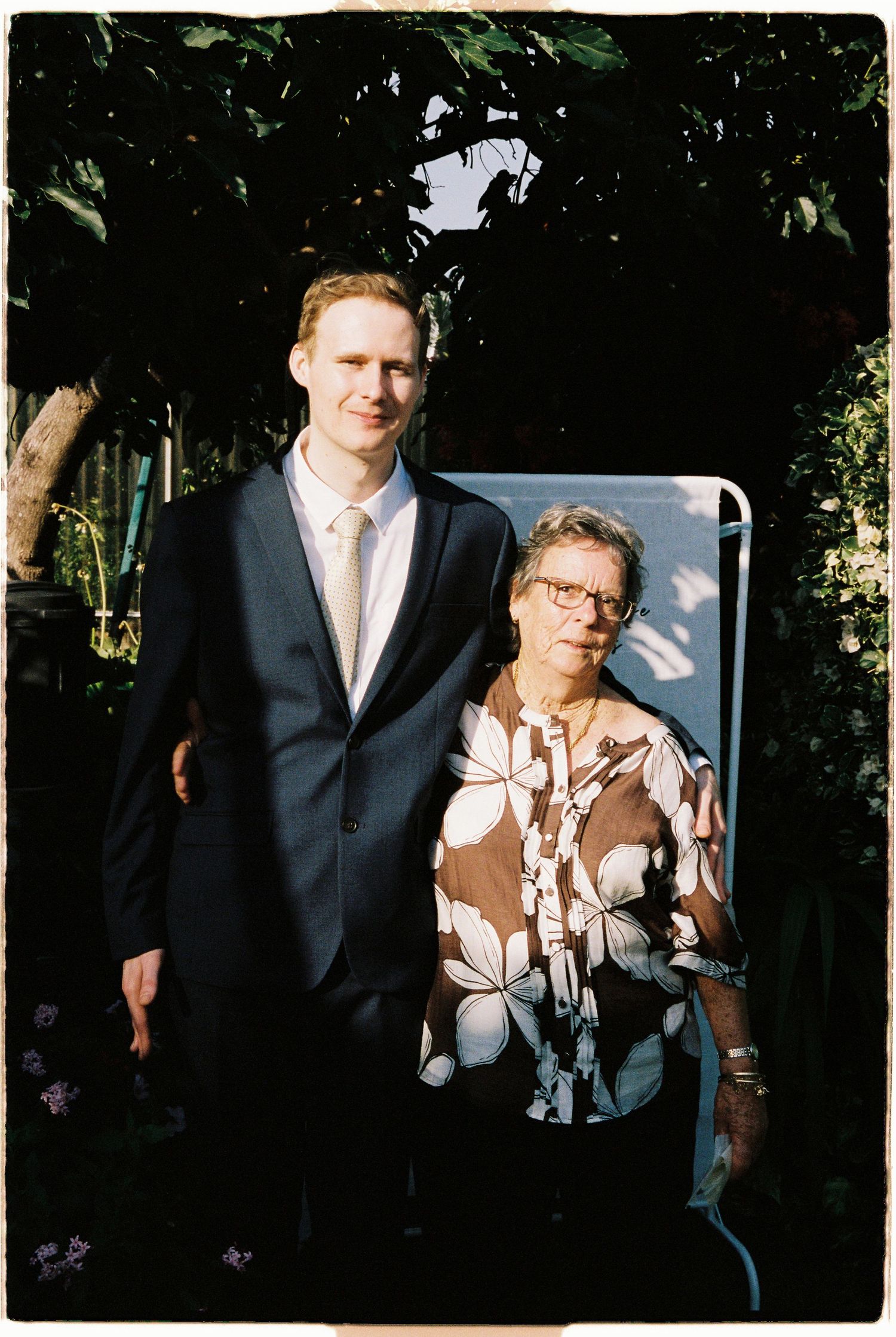 Tall groom in dark suit poses with elderly woman in floral blouse in a sunlit garden setting.