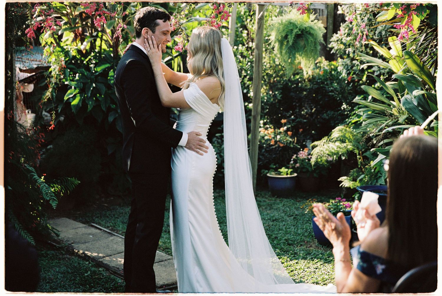 Bride and groom share first kiss at outdoor garden wedding ceremony while guest photographs the moment.