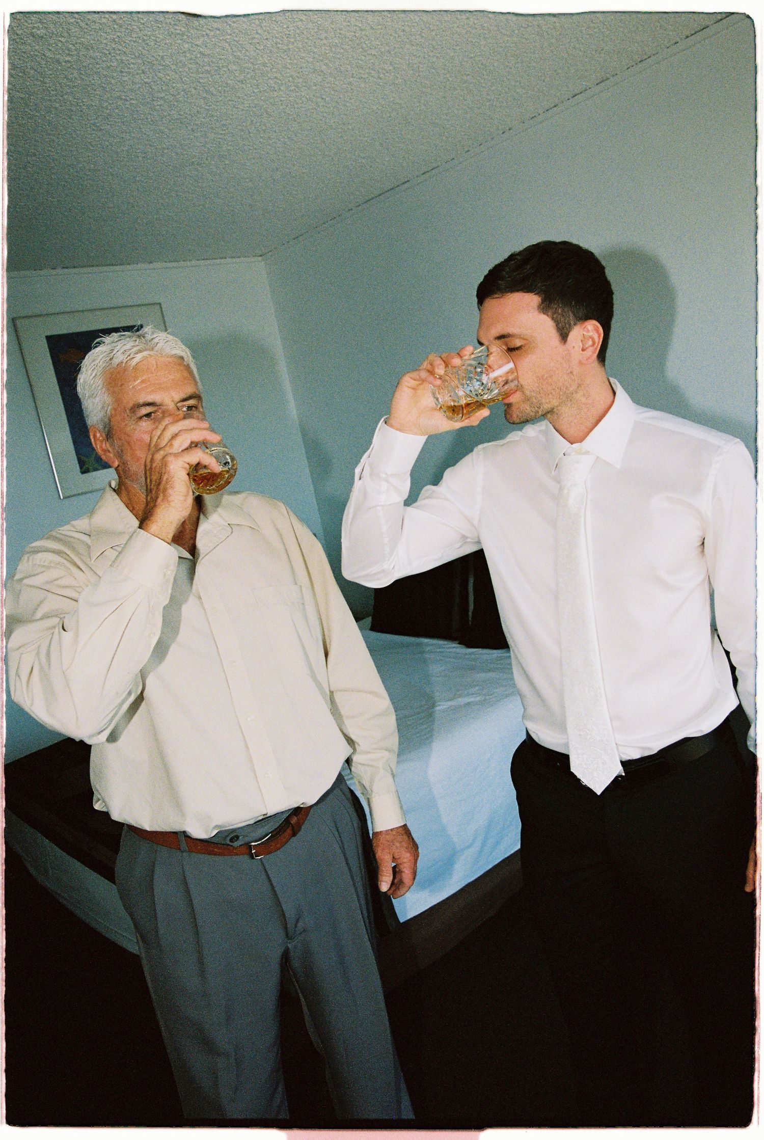 Older man and young groom drinking whiskey together in hotel room before wedding.