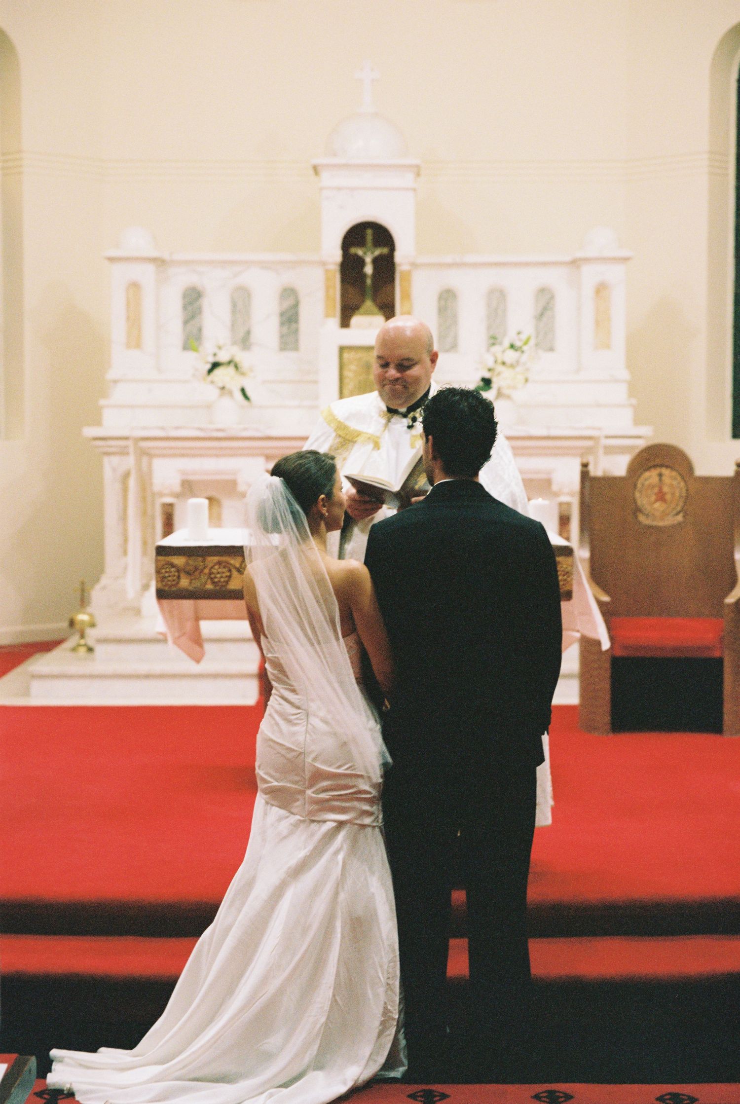 Bride and groom exchange vows before smiling officiant at ornate white church altar with red carpet.