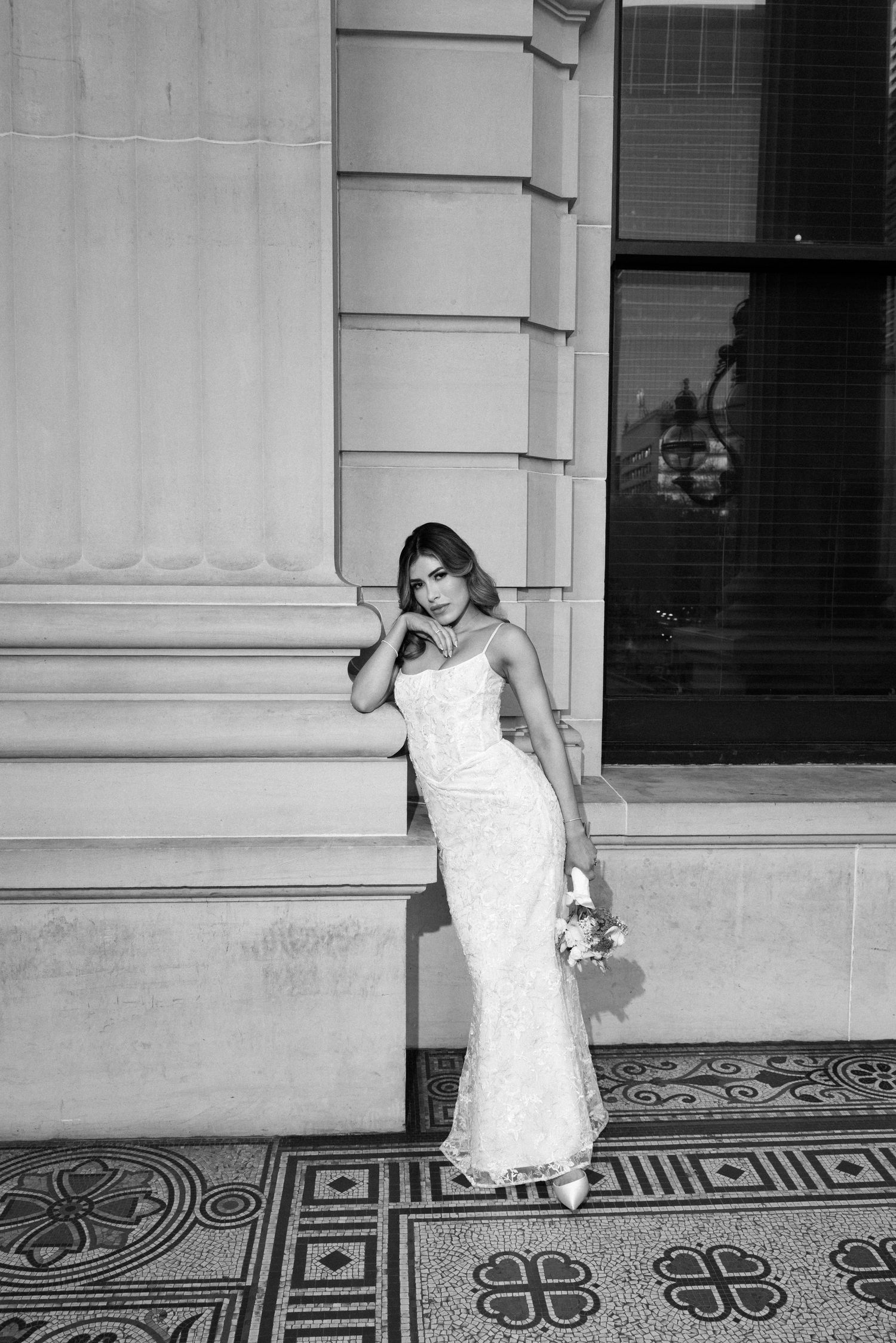 Black and white photo of a bride in a fitted lace gown holding flowers, posing against an elegant stone building.