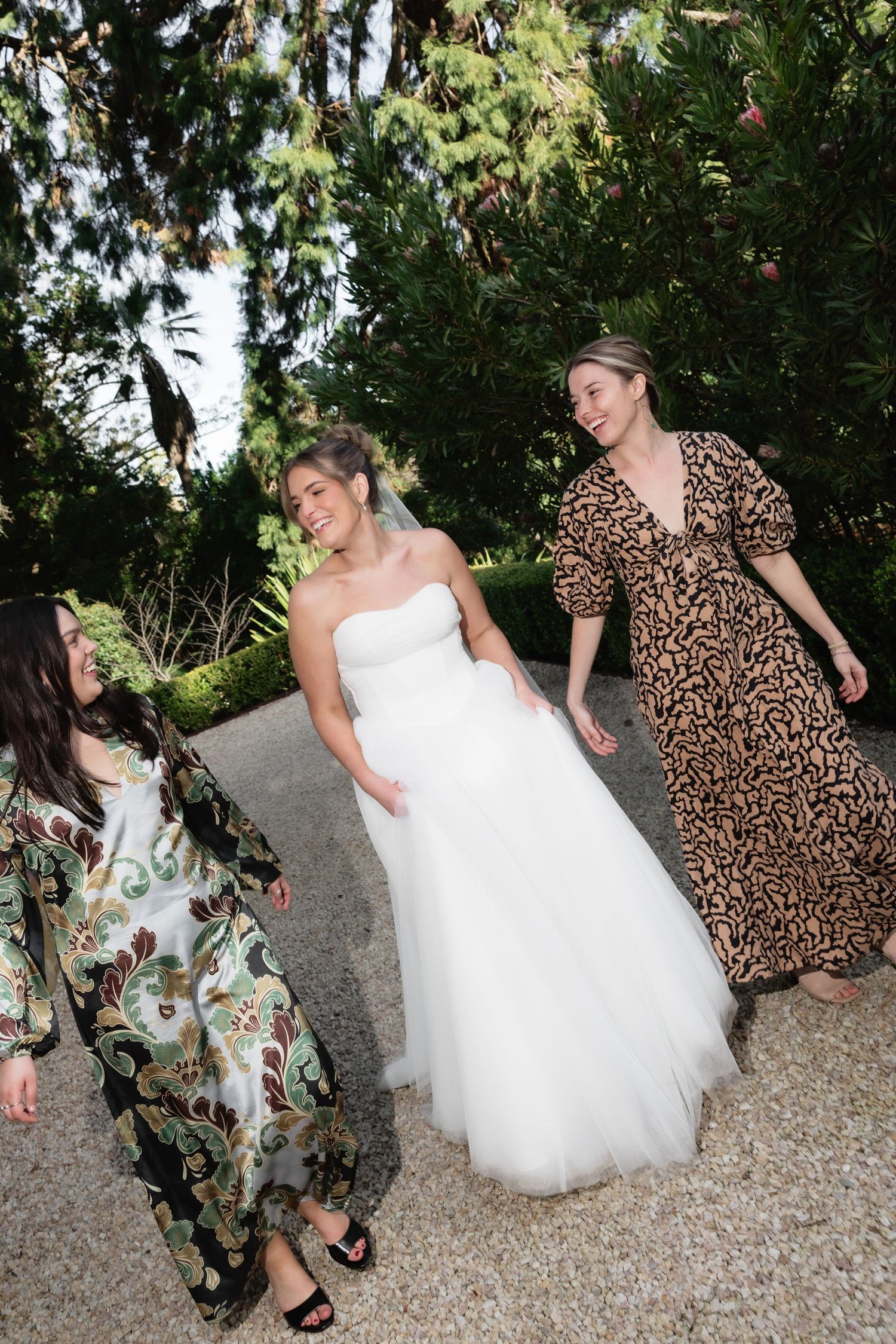 Bride in white tulle gown walking with two guests in garden setting.