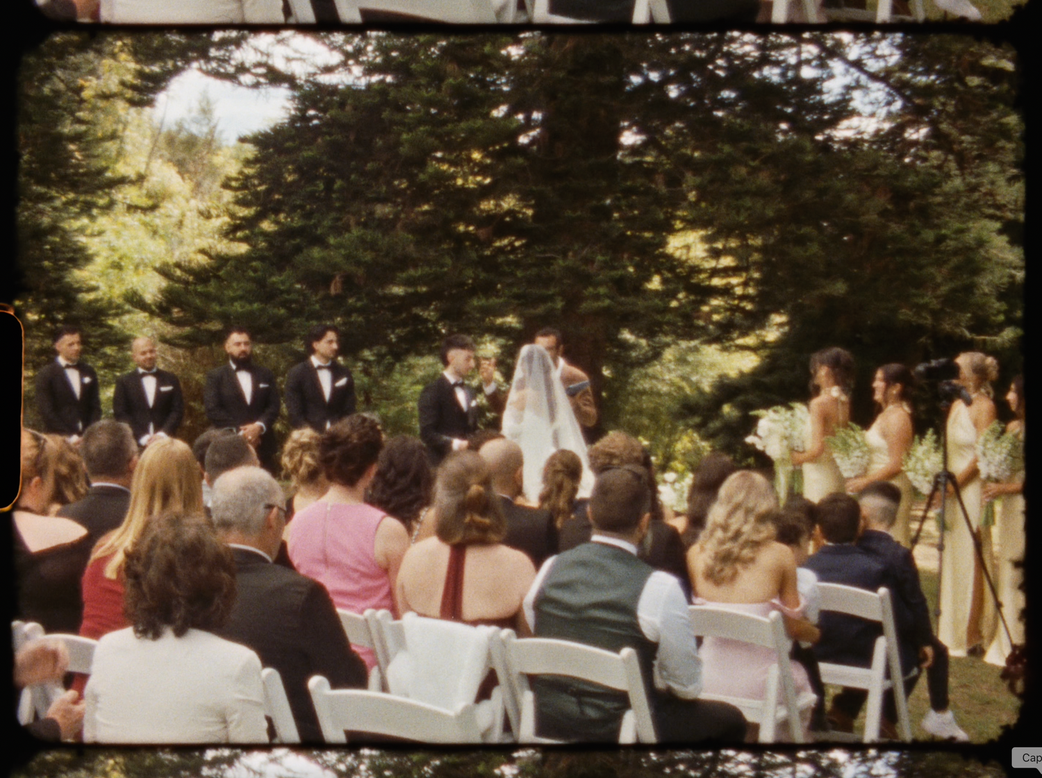 Outdoor wedding ceremony with guests seated on white chairs, wedding party at altar, surrounded by lush green trees.