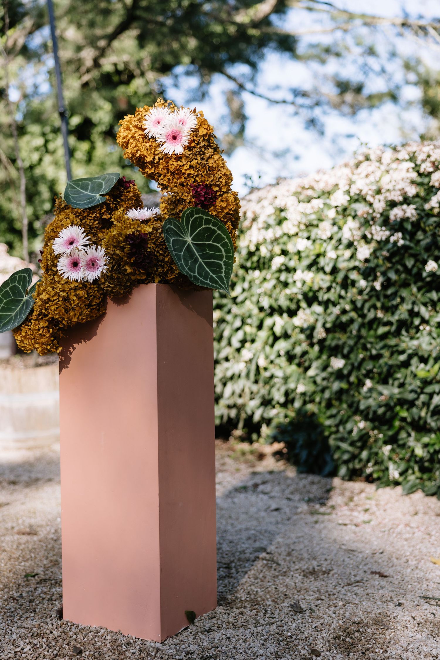 Pink pedestal vase with moss and pink gerbera daisy floral arrangement outdoors near a green hedge.