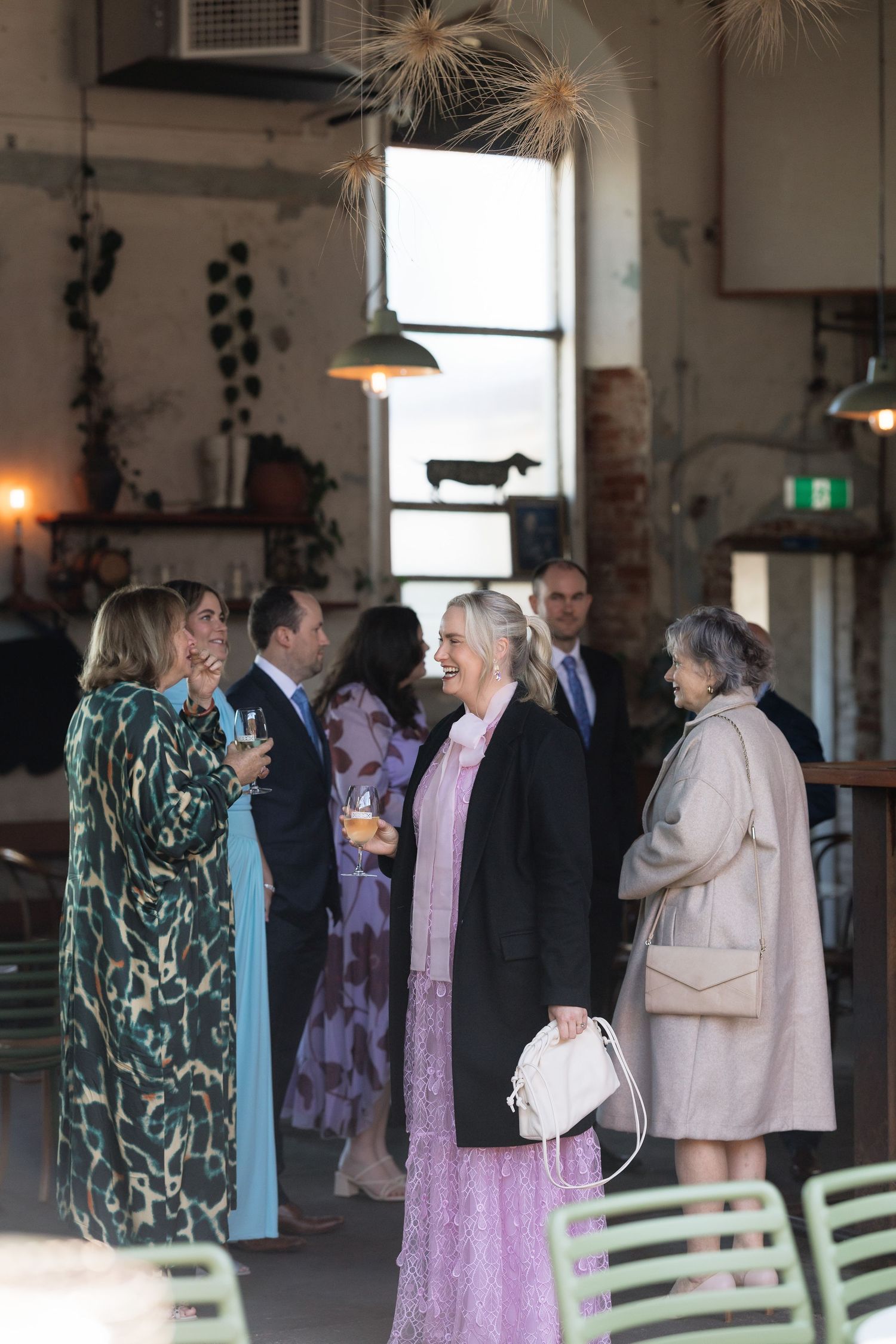 Guests mingling at an elegant indoor event, wearing formal attire in a rustic venue with pendant lights.