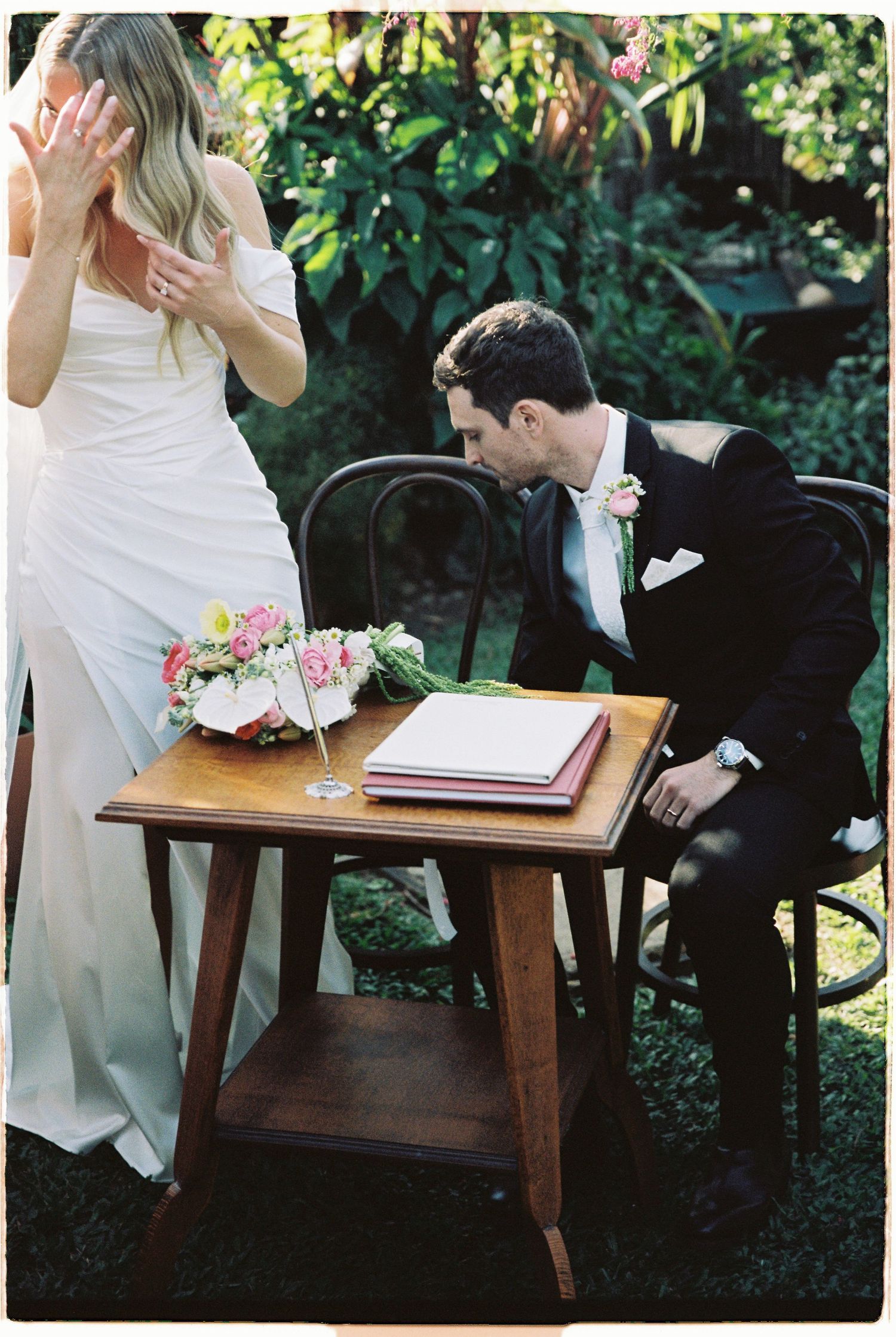 Groom in black tuxedo signing wedding registry at wooden table with pink floral bouquet beside bride in white gown.