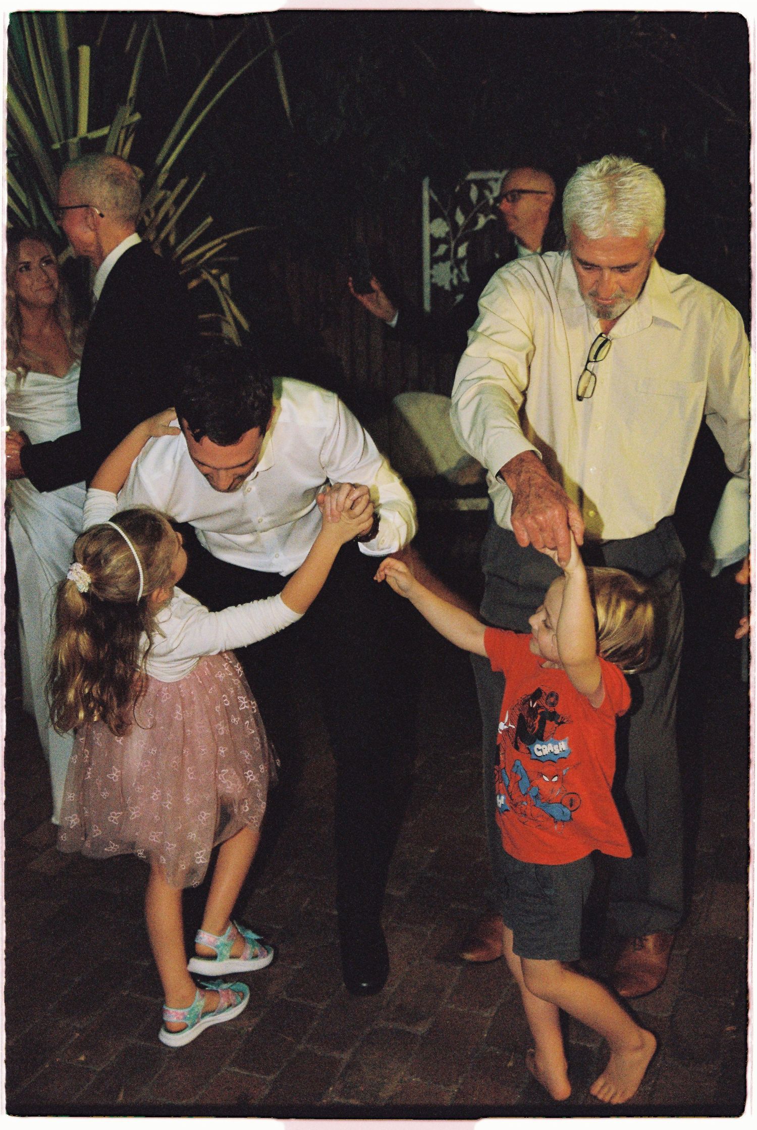 Two men dancing with young children at an outdoor evening wedding reception.